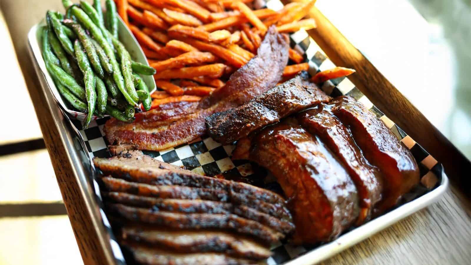 A tray of Brileys BBQ delights featuring sliced brisket, ribs, bacon, sweet potato fries, and green beans served on a checkered paper liner.