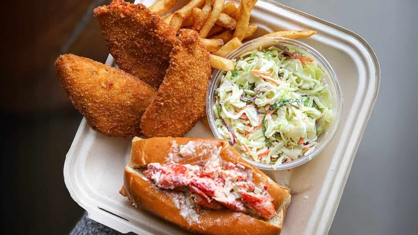A takeout container from Counter Culture: Burien Fish House features crispy fried fish fillets, golden French fries, creamy coleslaw, and a lobster roll on a split-top bun.