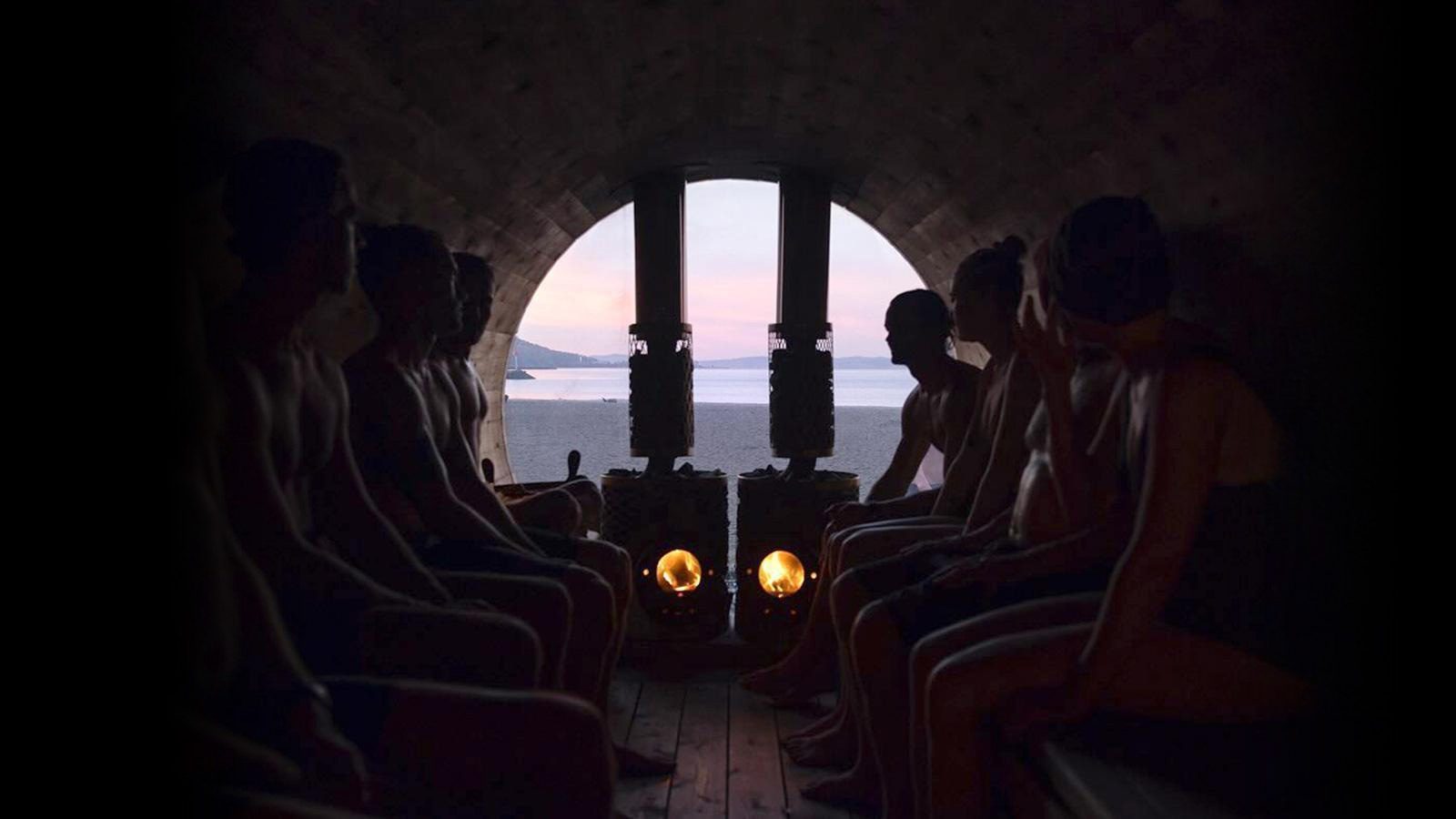 Inside a dark sauna, people bask in the warmth, gazing toward an open door that frames a breathtaking view of a sunset over the beach and sea. It feels like a serene escape, much like experiencing "A Seattle New Year For All Types," where everyone finds their own moment of peace and renewal.