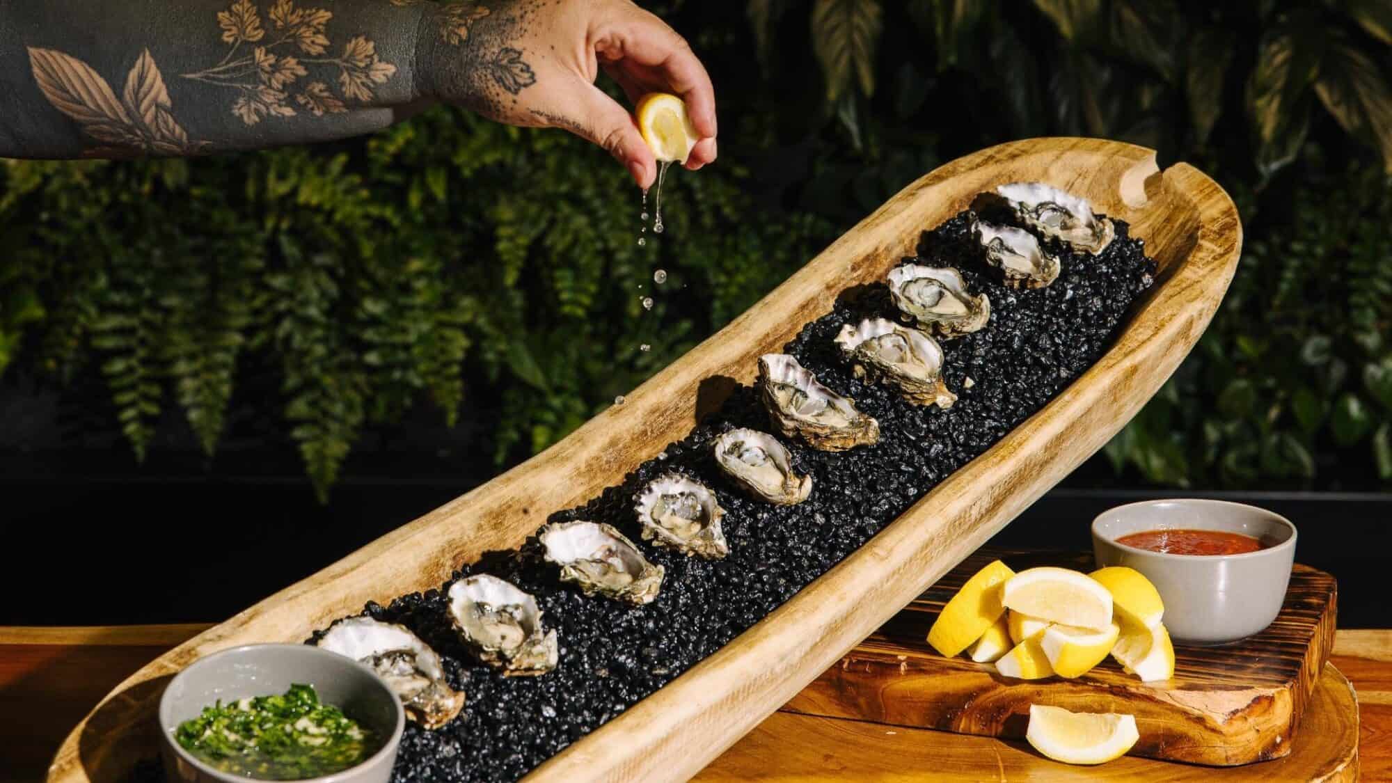 A hand squeezes lemon over a long wooden platter of raw oysters on black stones, with bowls of green sauce, cocktail sauce, and lemon wedges beside it.