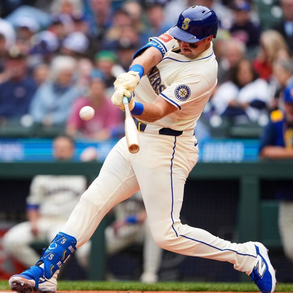 Mariners baseball player in a white uniform swings a bat and makes contact with the ball during a playoffs game, with spectators visible in the background.