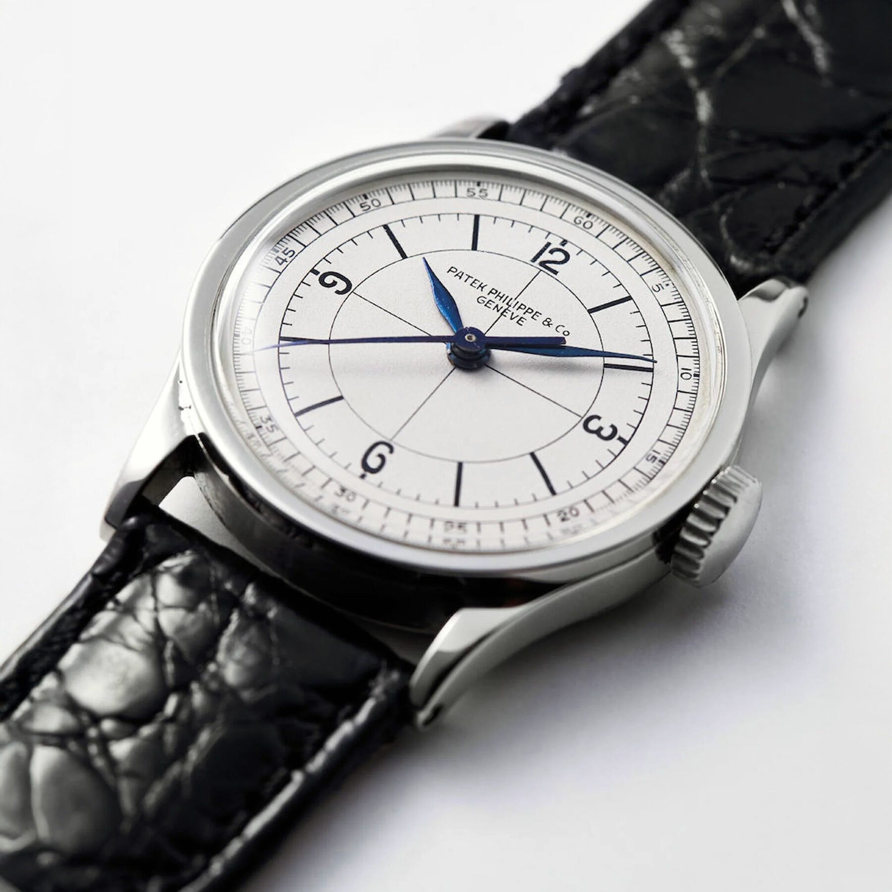 Close-up of a wristwatch with a white dial, blue hands, black numerals, and a textured black leather strap—an example of timeless product design. The dial reads "Patek Philippe Genève & Co.