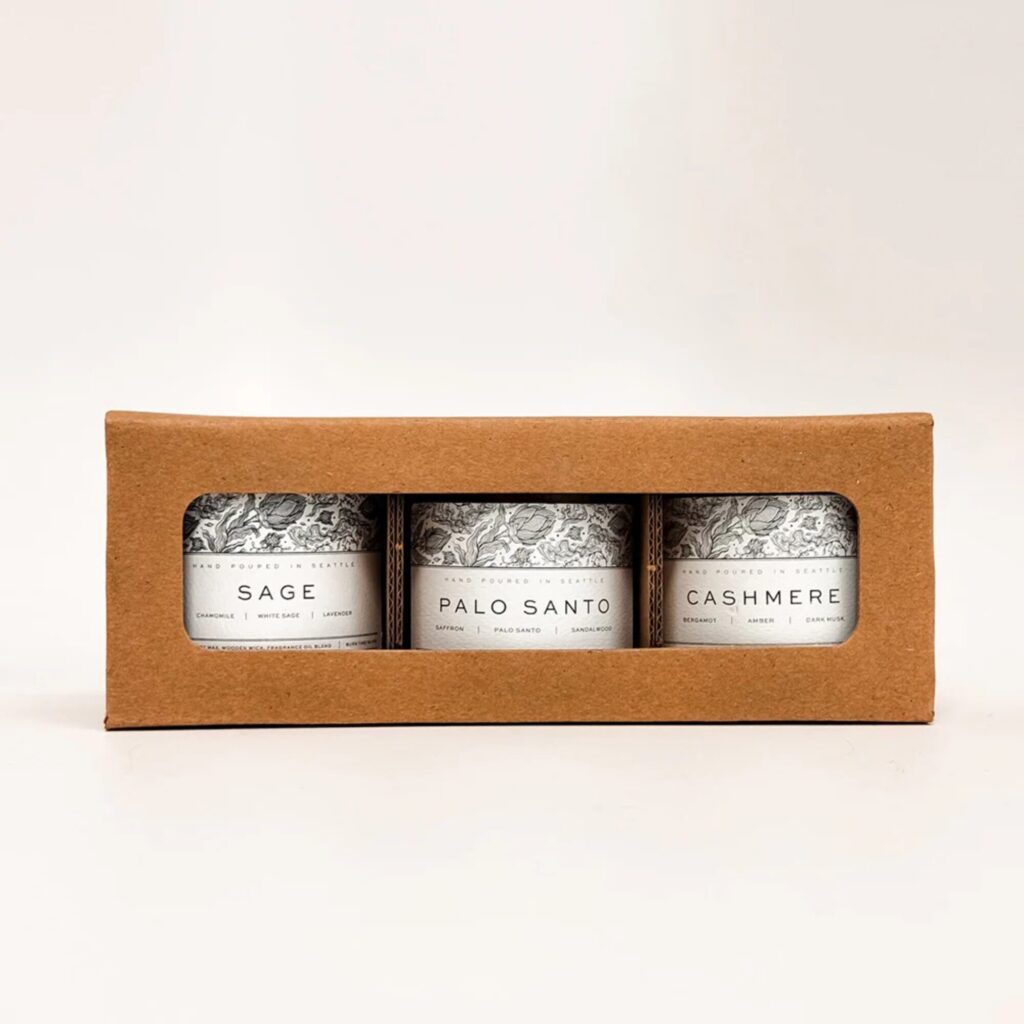 A brown cardboard box with a clear window displays three candles labeled Sage, Palo Santo, and Cashmere.