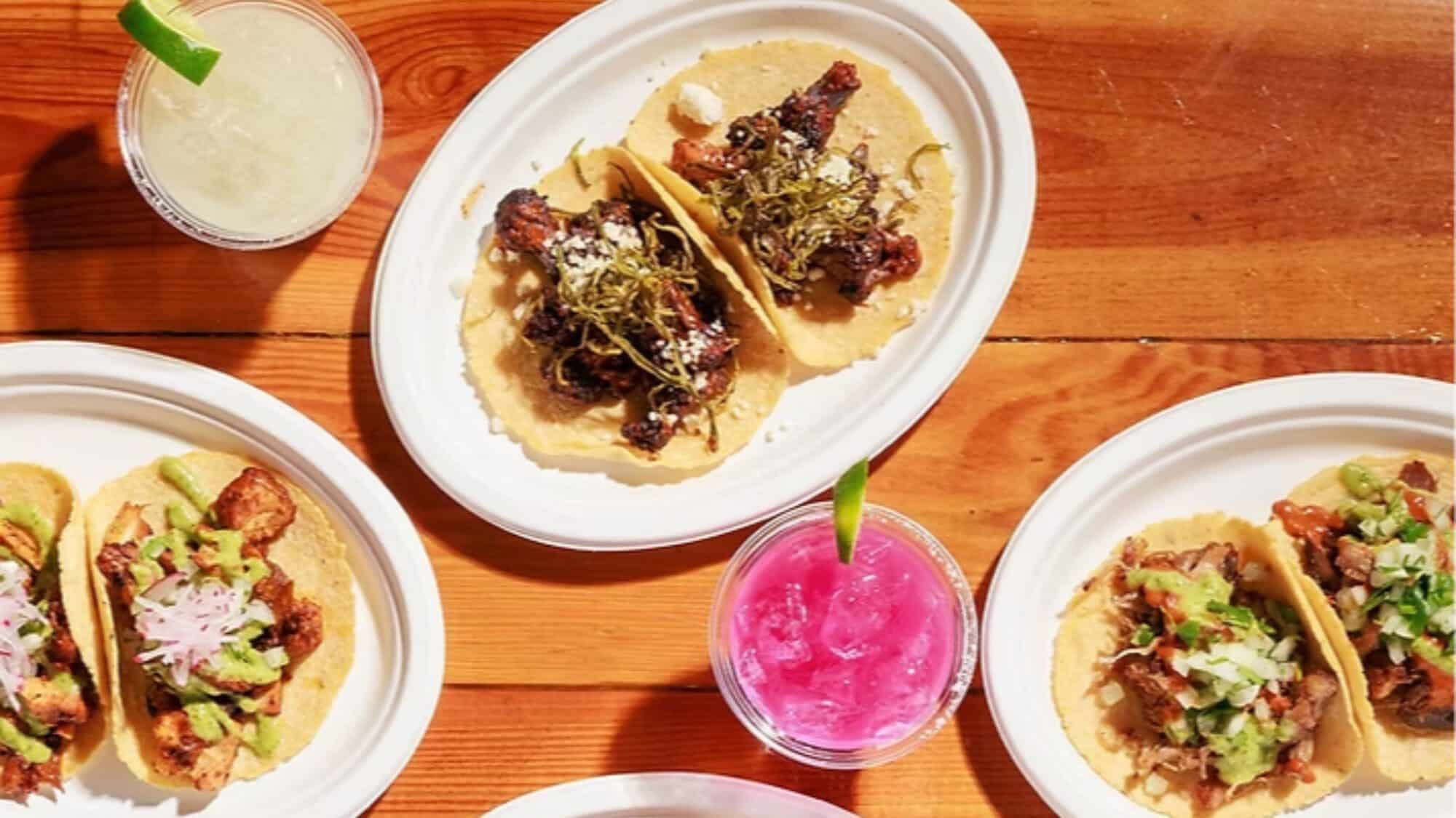 Three plates of tacos with assorted toppings and two colorful drinks on a wooden table.