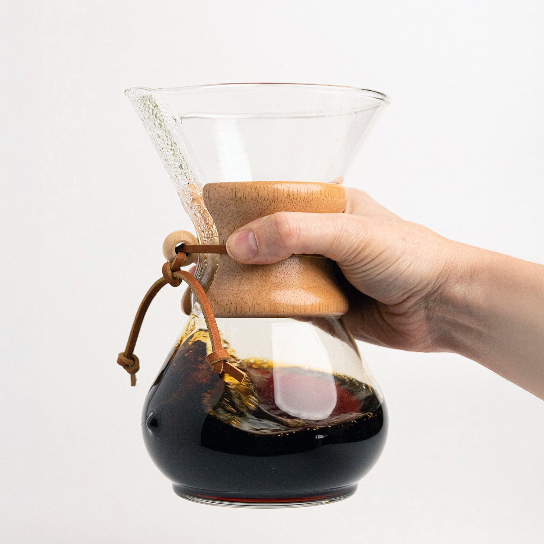 A hand holding a glass Chemex coffee maker with brewed coffee inside, against a plain white background, celebrates the simple beauty of design around us.