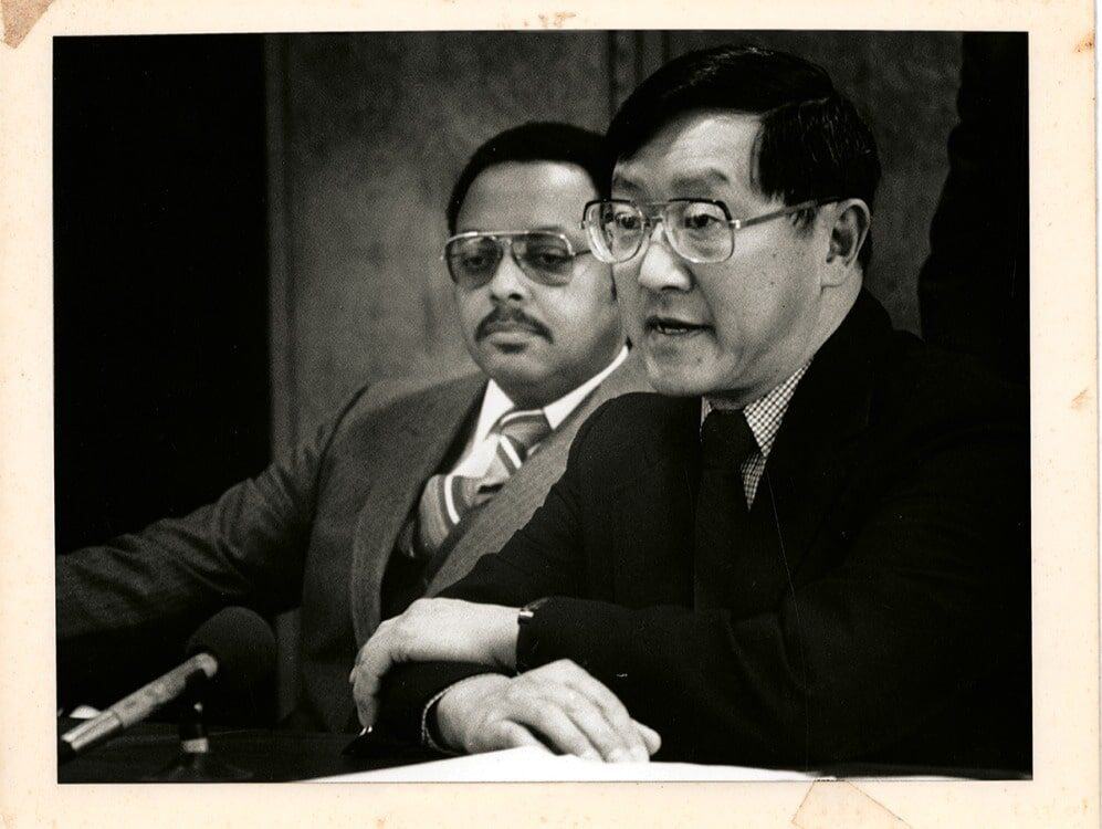 Two men in suits sit at a table with microphones, one speaking about resilience while the other listens attentively in the background.