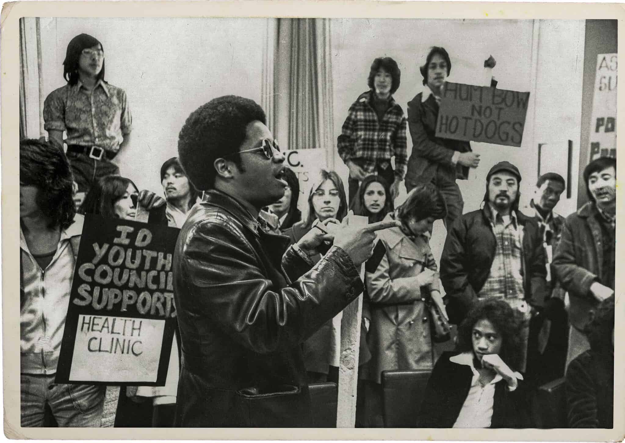 A group of people, some holding protest signs, gather indoors; one man in sunglasses and a leather jacket speaks about resilience while others listen or hold signs reflecting their resistance.
