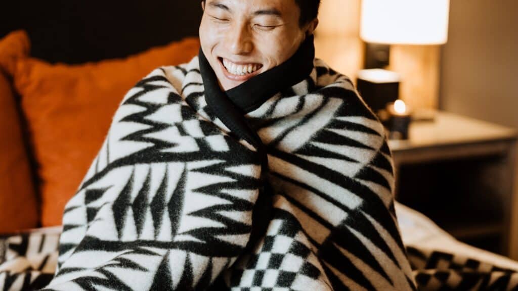 A person smiles warmly while wrapped in a patterned black and white blanket, sitting indoors with a soft light in the background, celebrating Native American Heritage Month.