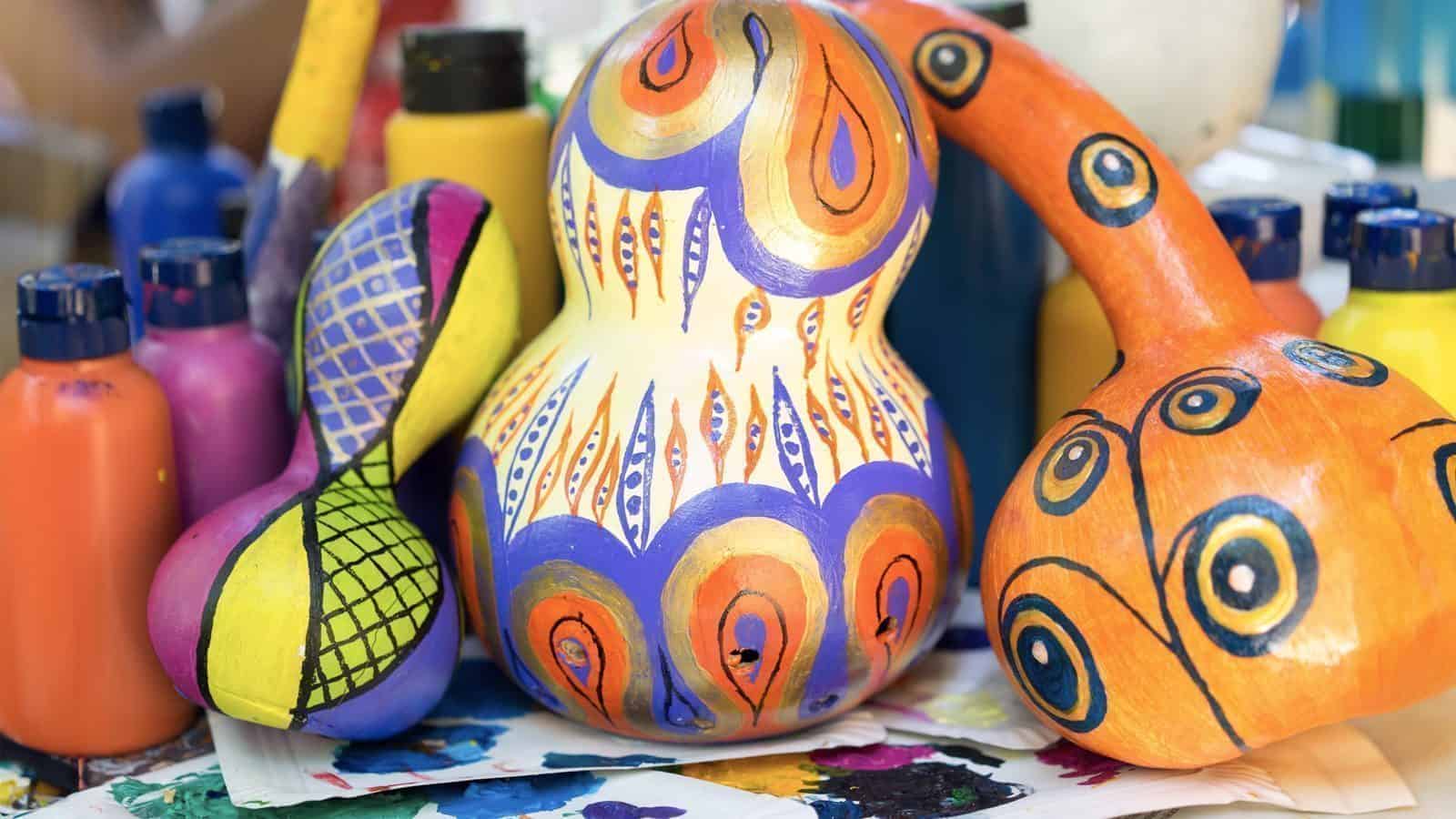 The workspace brims with curiosity, as vibrantly painted gourds adorned with abstract patterns are surrounded by paint bottles, inviting endless possibilities.