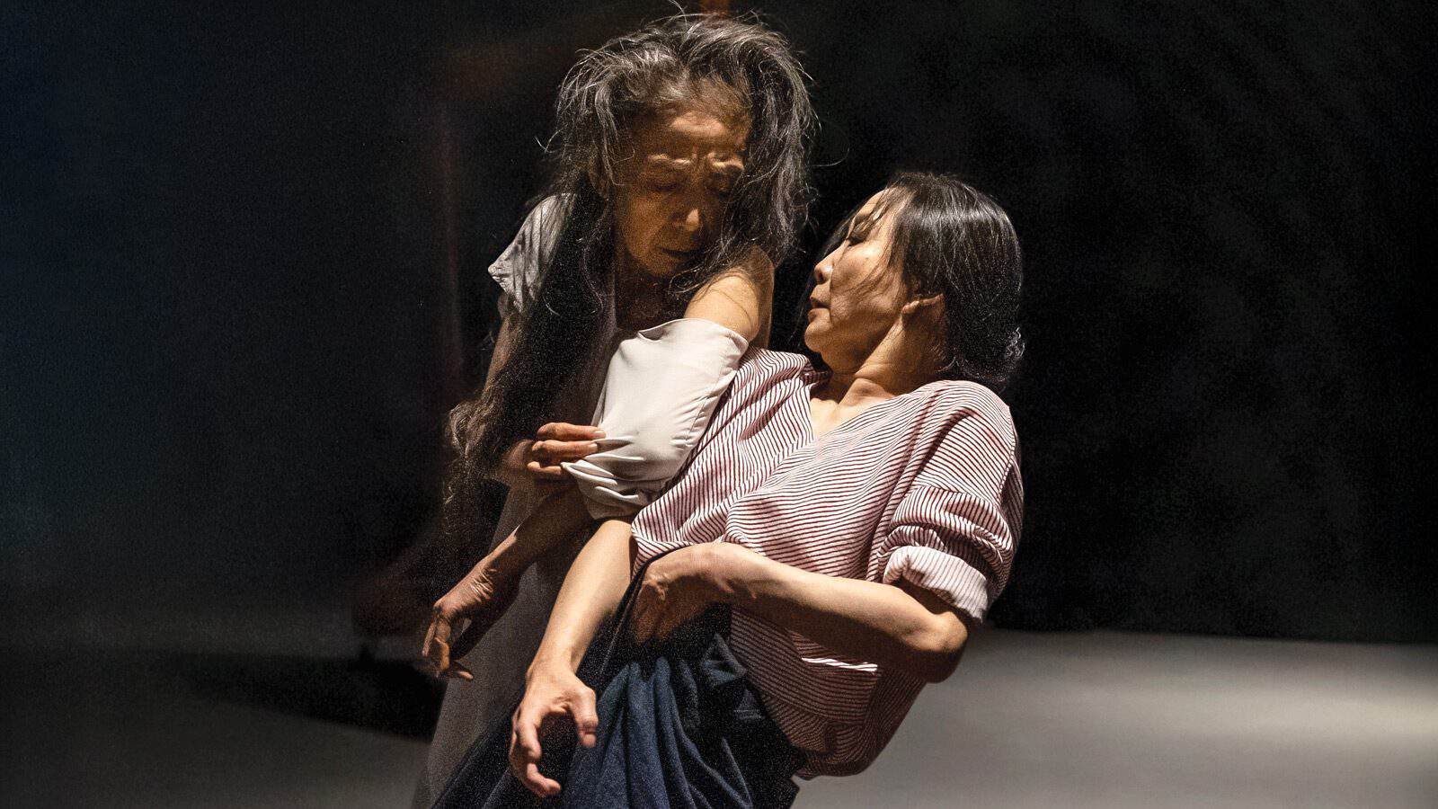 Two women with long dark hair perform an expressive dance, one leaning forward and supporting the other, both in loose clothing on a dimly lit stage—an evocative glimpse into Seattle's vibrant autumn arts scene.