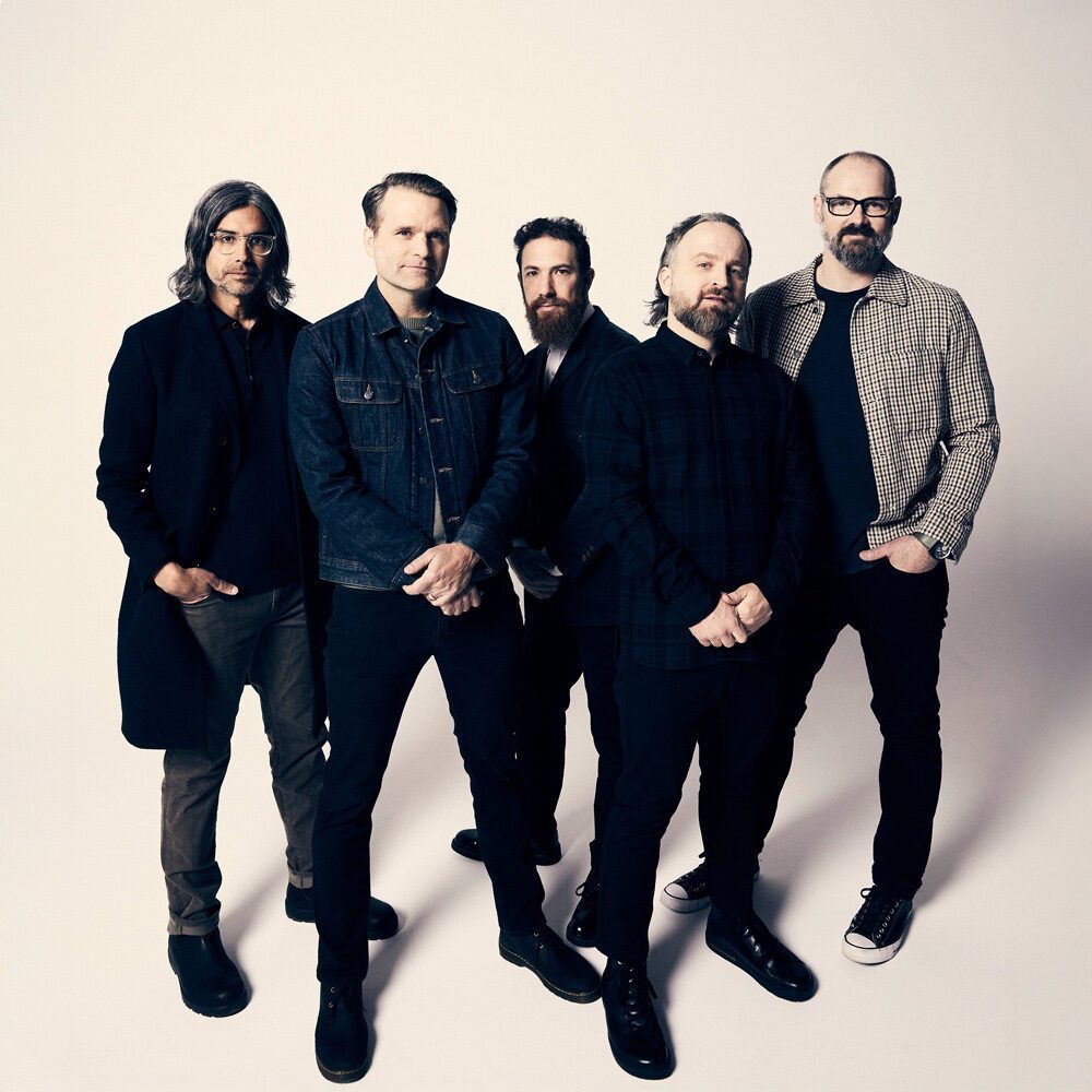 Five men stand together against a plain white background, dressed in casual and semi-formal clothing, facing the camera—a Must List shot to remember on 07-31-2025.