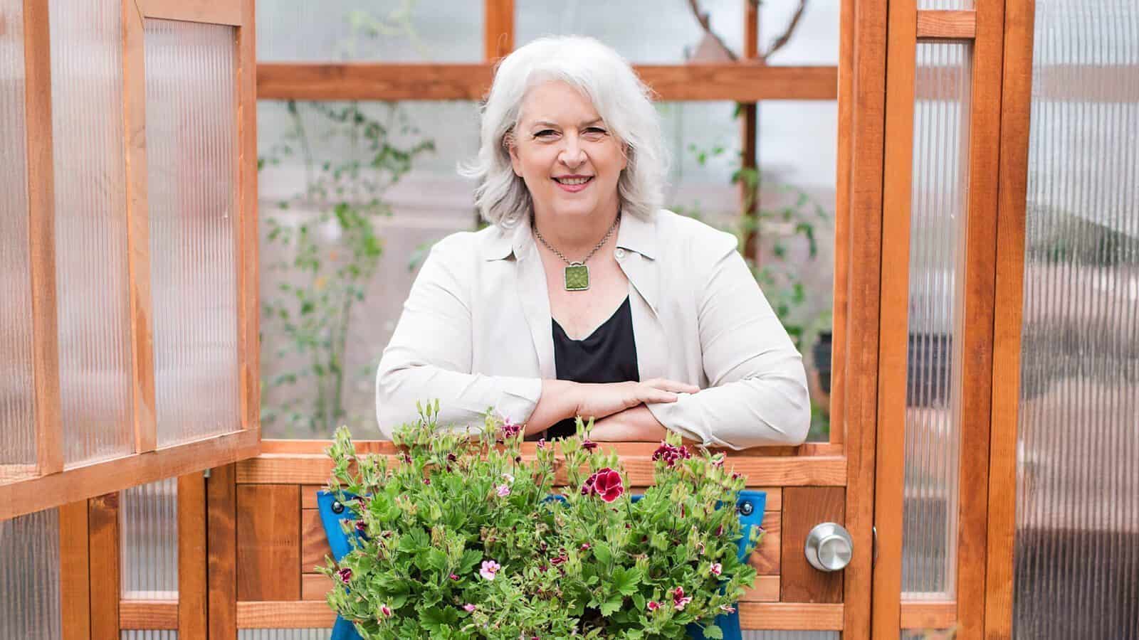 An older woman with gray hair stands smiling behind a wooden door, with blooming local flowers in front of her inside a greenhouse—a scene reminiscent of Debra Prinzing's focus on seasonal and sustainably harvested flowers.