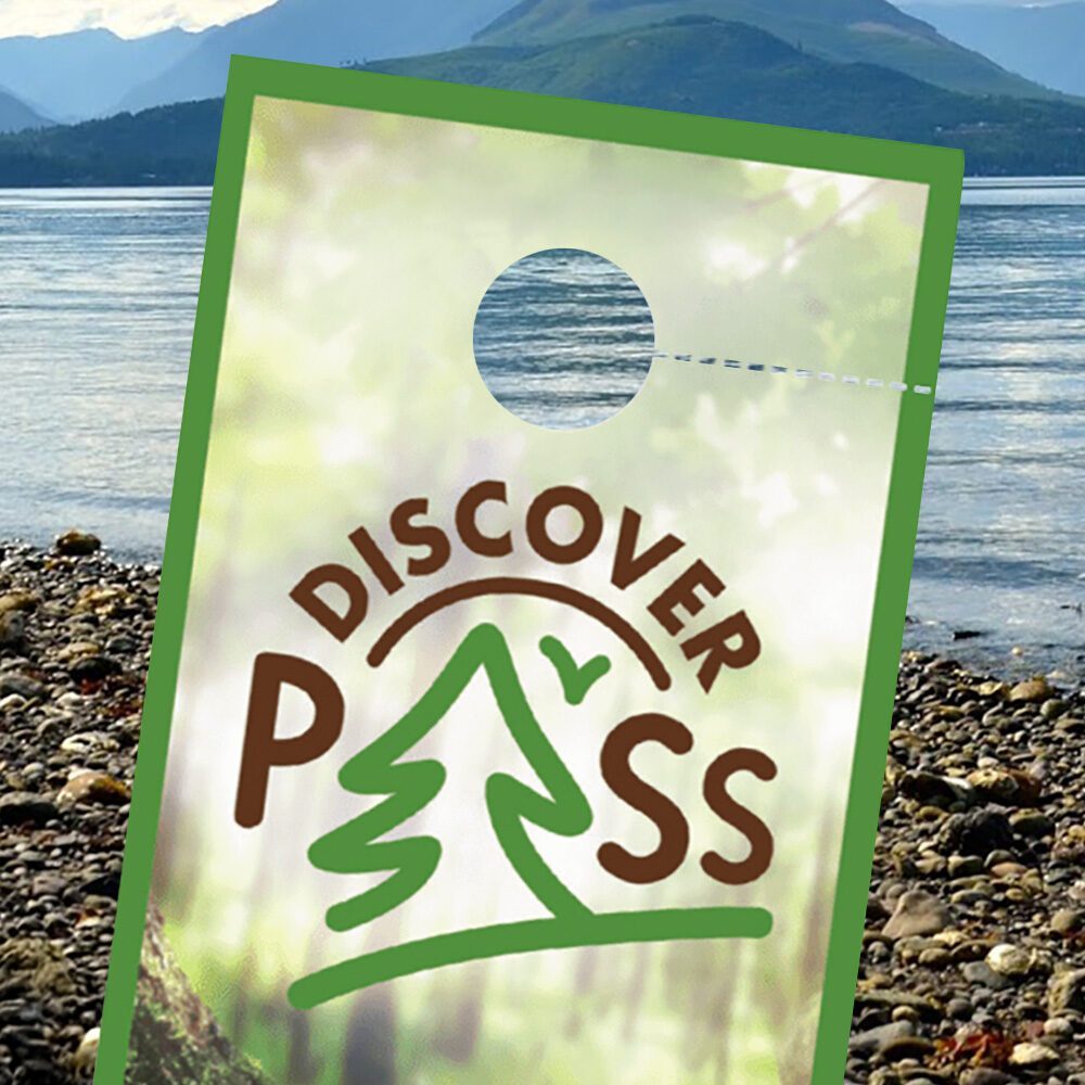 A Discover Pass, valid through 08-07-2025, hangs in front of a rocky lakeshore with mountains and trees in the background on a clear day—a Must List scene for outdoor adventurers.