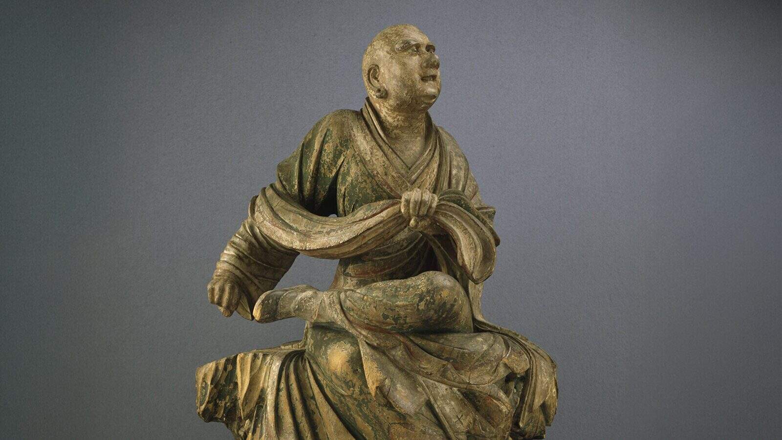 A wooden statue of a seated monk with one leg crossed, wearing flowing robes and holding a section of the garment, evokes the rich heritage seen in Seattle’s Cultural Landmarks and honors the traditions of the AANHPI Community.