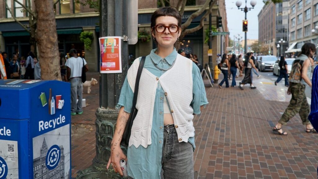 Person standing on a city sidewalk wearing glasses, a blue shirt, a white vest, and gray jeans, with people and buildings visible in the background.