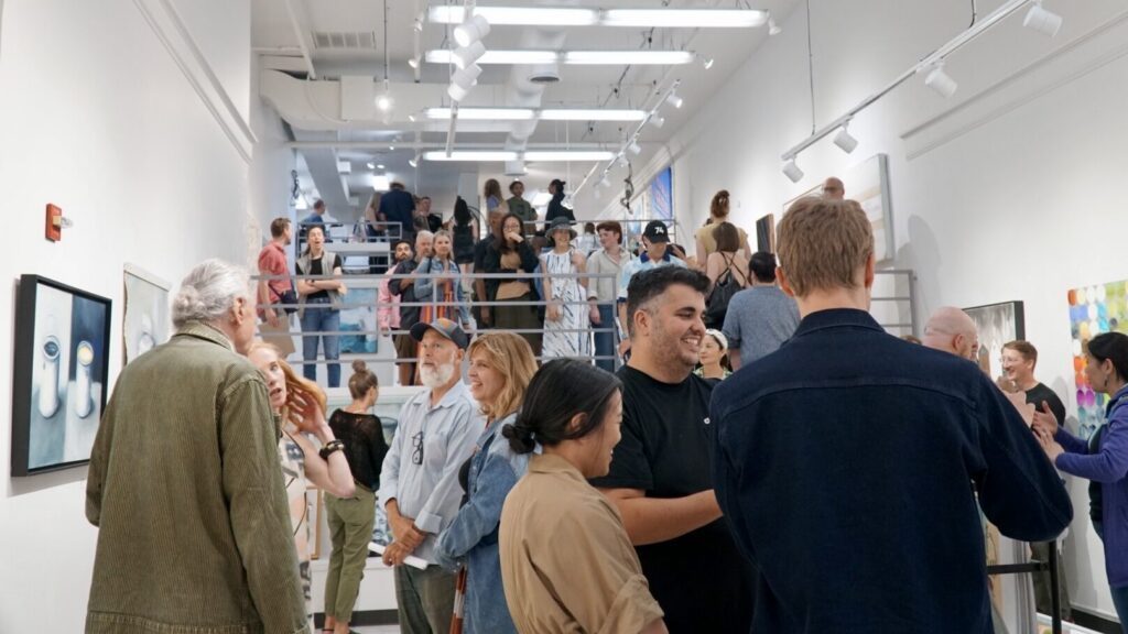 A group of people attend an art gallery event, viewing artwork on the walls and conversing in a brightly lit, modern space.