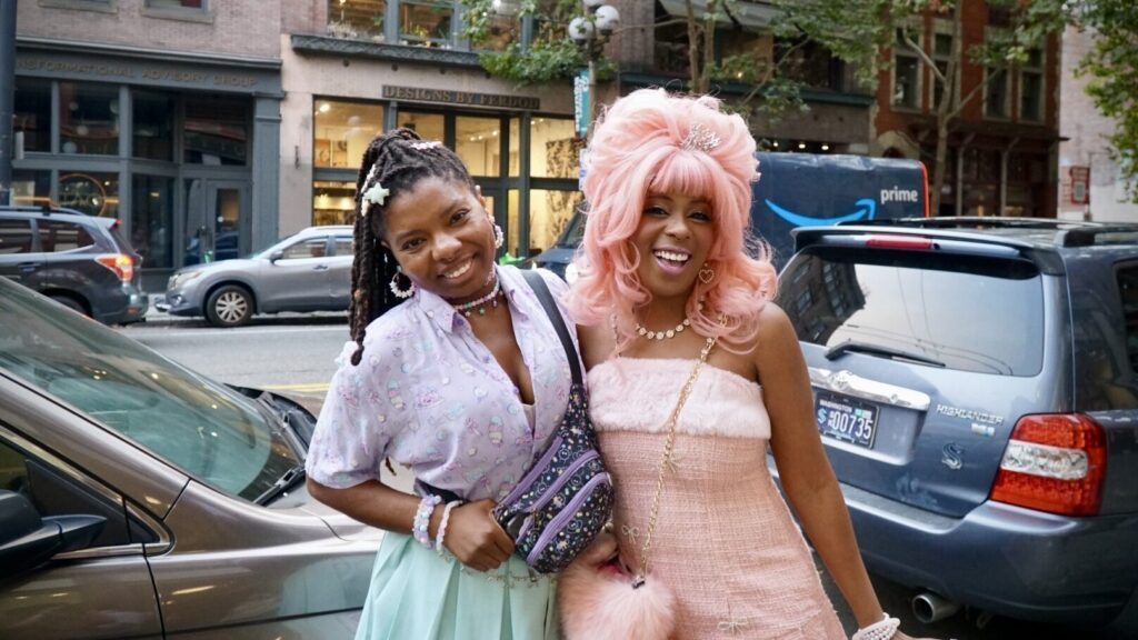Two women stand smiling on a city street, wearing colorful and stylish outfits; one in pastel purple and green, the other in a pink dress with a large pink wig. Cars and buildings are in the background.