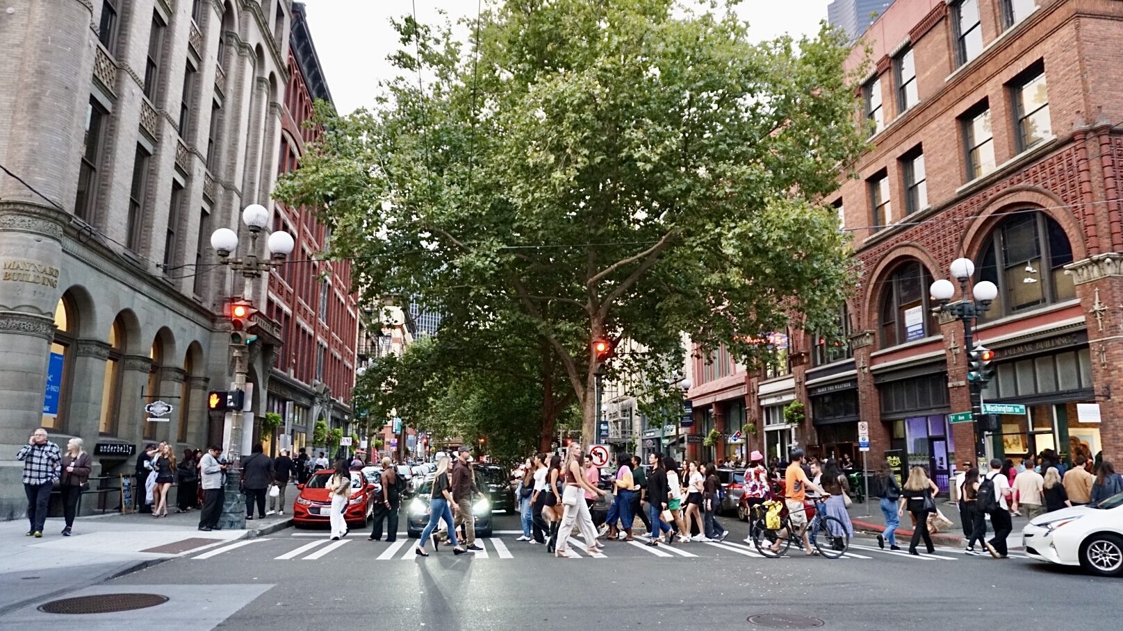 A busy city intersection with many pedestrians crossing the street, surrounded by historic brick buildings and large leafy trees.