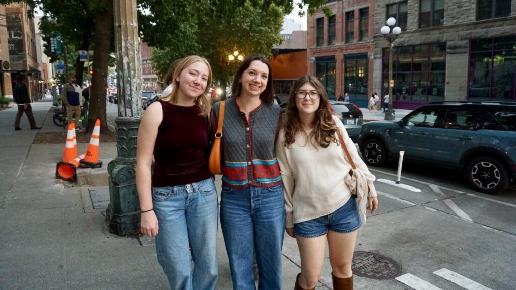 Three young women stand together and smile on a city sidewalk near parked cars and buildings, with trees and street cones visible in the background.