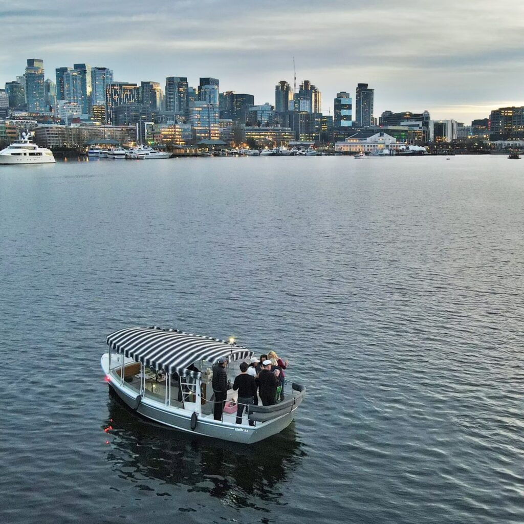 A small group of people stands on a covered boat in a body of water with a city skyline and buildings in the background.