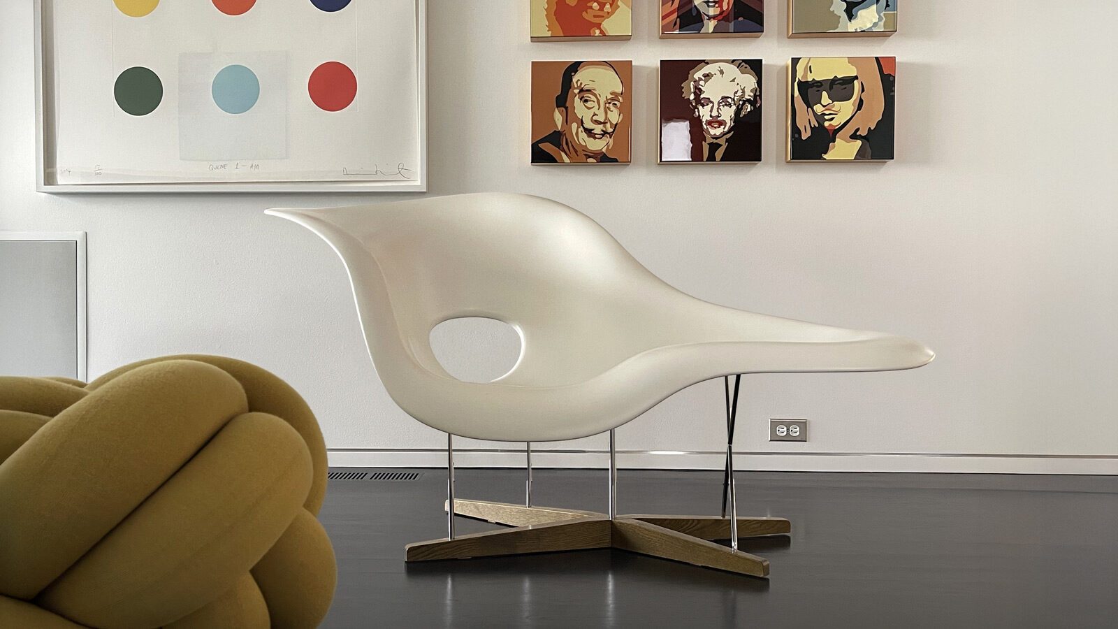 A modern white sculptural chair, reminiscent of the iconic Eames La Chaise, sits in a minimalist room with colorful artwork and portraits displayed on a white wall, highlighting the beauty of design around us.