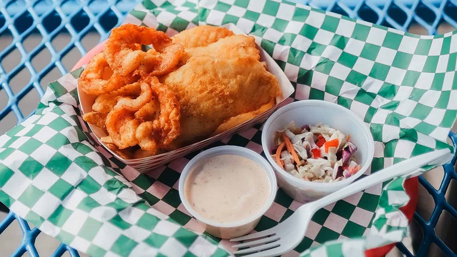 A paper tray holds fried fish and chips, onion rings, a small cup of coleslaw, and spicy tartar sauce, placed on a green checkered liner atop a blue metal table.