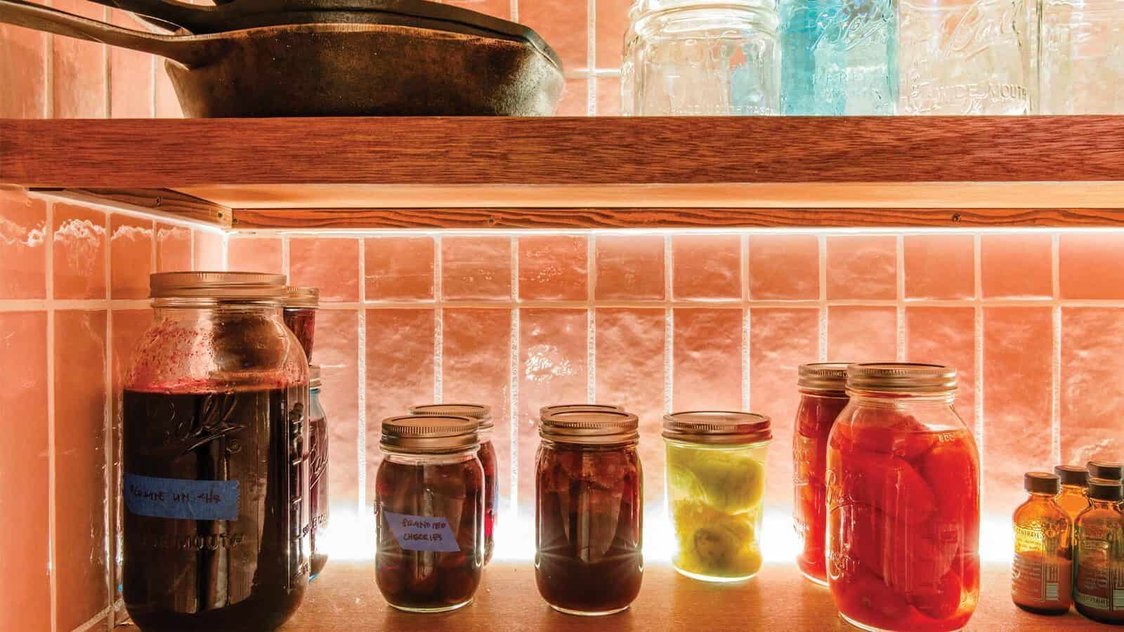 Jars of preserved fruits and vegetables sit on wooden shelves in front of pink-tiled walls, echoing a Frank Lloyd Wright-inspired aesthetic, with a dark pot and glass containers above.