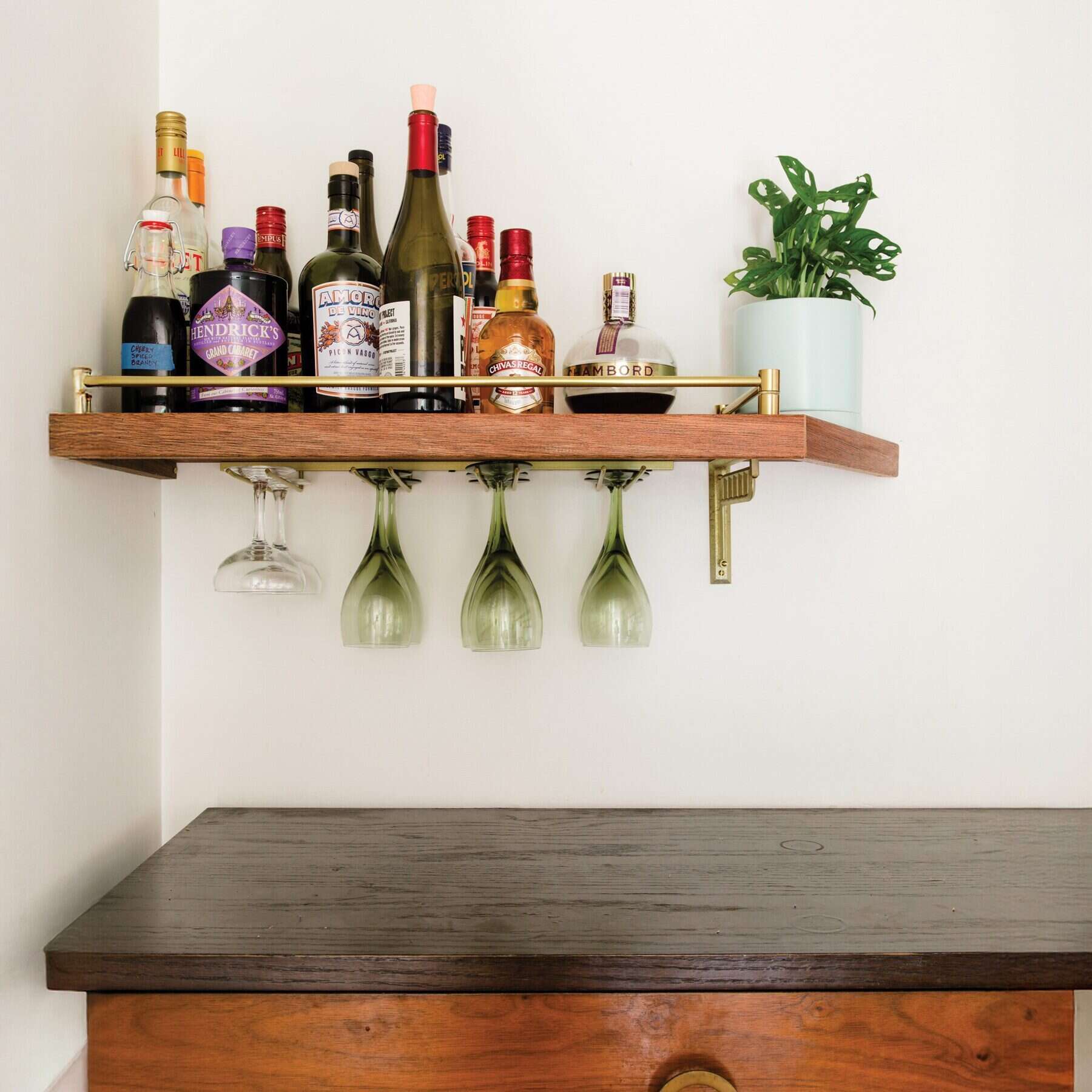 A wooden shelf with various liquor bottles and a potted plant exudes Frank Lloyd Wright's influence. Below, four wine glasses hang upside down, while a wooden table, reminiscent of home renovations by architects, completes the setup.