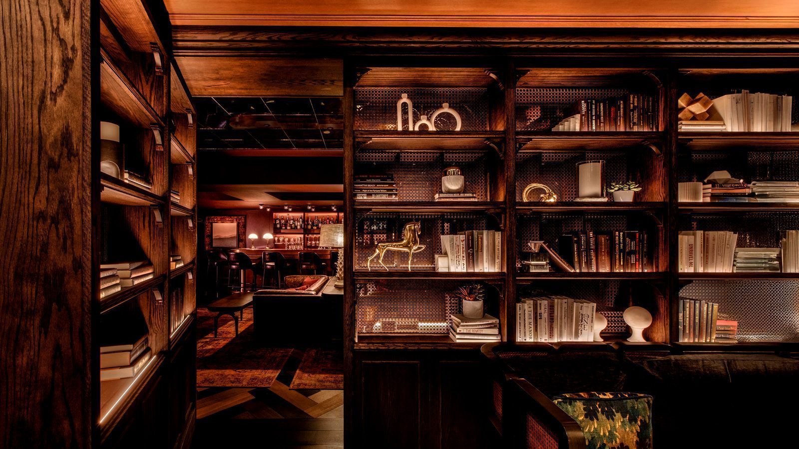 In a Seattle New Year setting, this dimly lit cozy library boasts wooden bookshelves brimming with books and decorative treasures, perfect for bibliophiles of all types. A glimpse of the bar area in the background invites guests to celebrate and unwind.