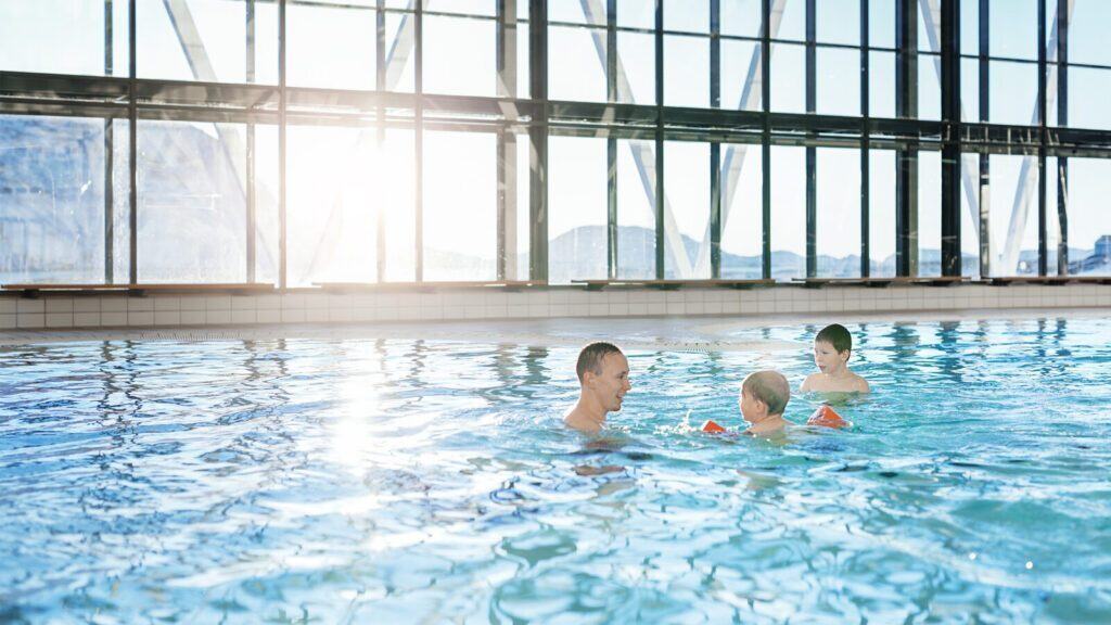 Three people are swimming in an indoor pool with large windows letting in sunlight. Two children wear orange floaties, and an adult is with them in the water.
