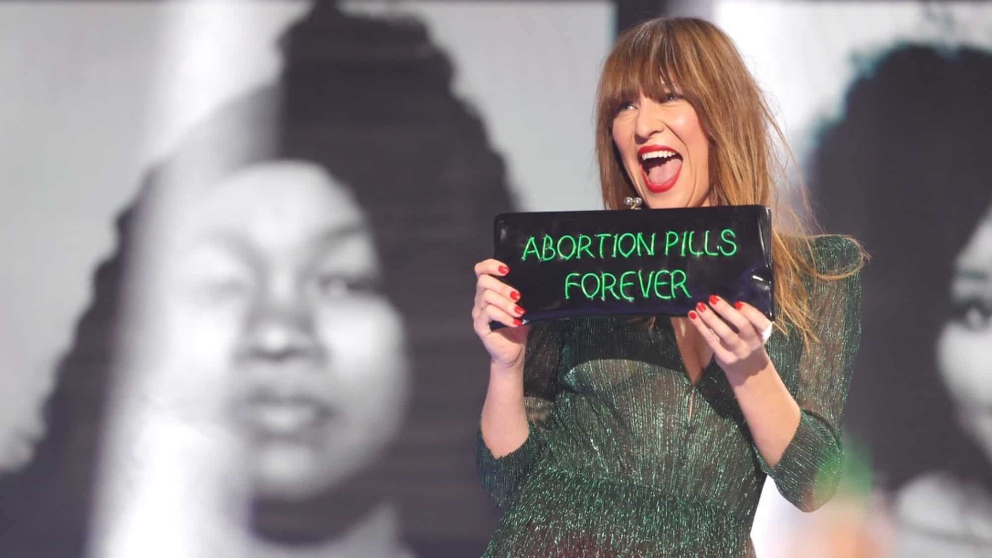 A woman in a green dress holds a black sign with green writing that reads "ABORTION PILLS FOREVER," standing in front of black-and-white portrait images.