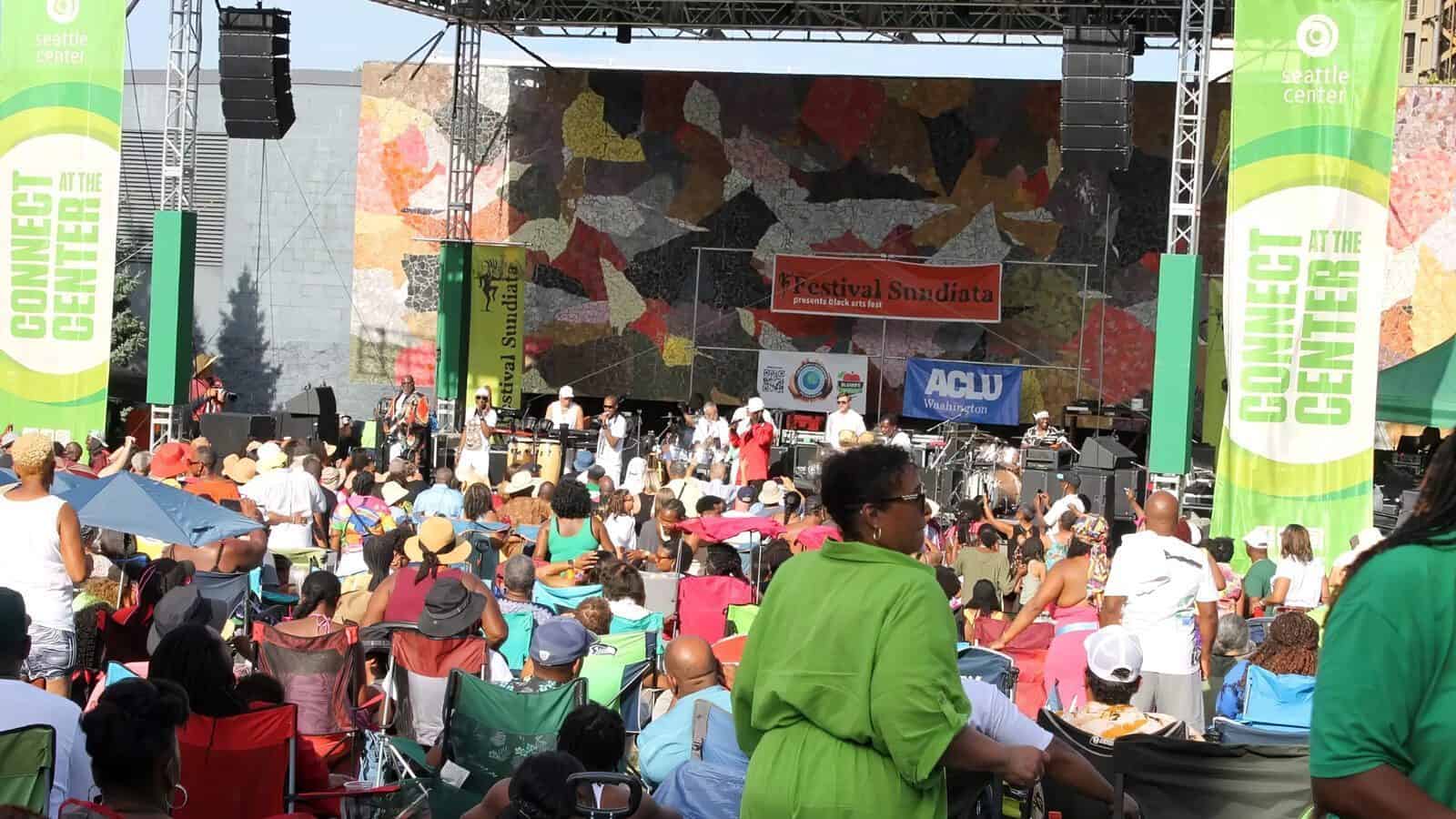 A crowd sits in front of an outdoor stage at Festival Sundiata, a must list event with banners displaying “Connect Center” and “Seattle Center” on both sides.