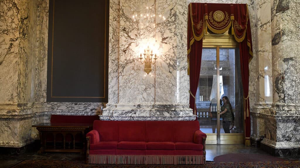 A red velvet couch sits against a marble wall beneath an ornate light fixture; a person is seen through glass doors framed by red and gold curtains.
