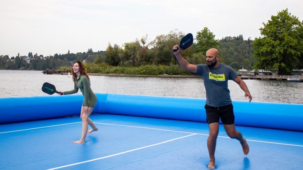Two people play pickleball barefoot on an inflatable blue court near a lake, with trees and water in the background.