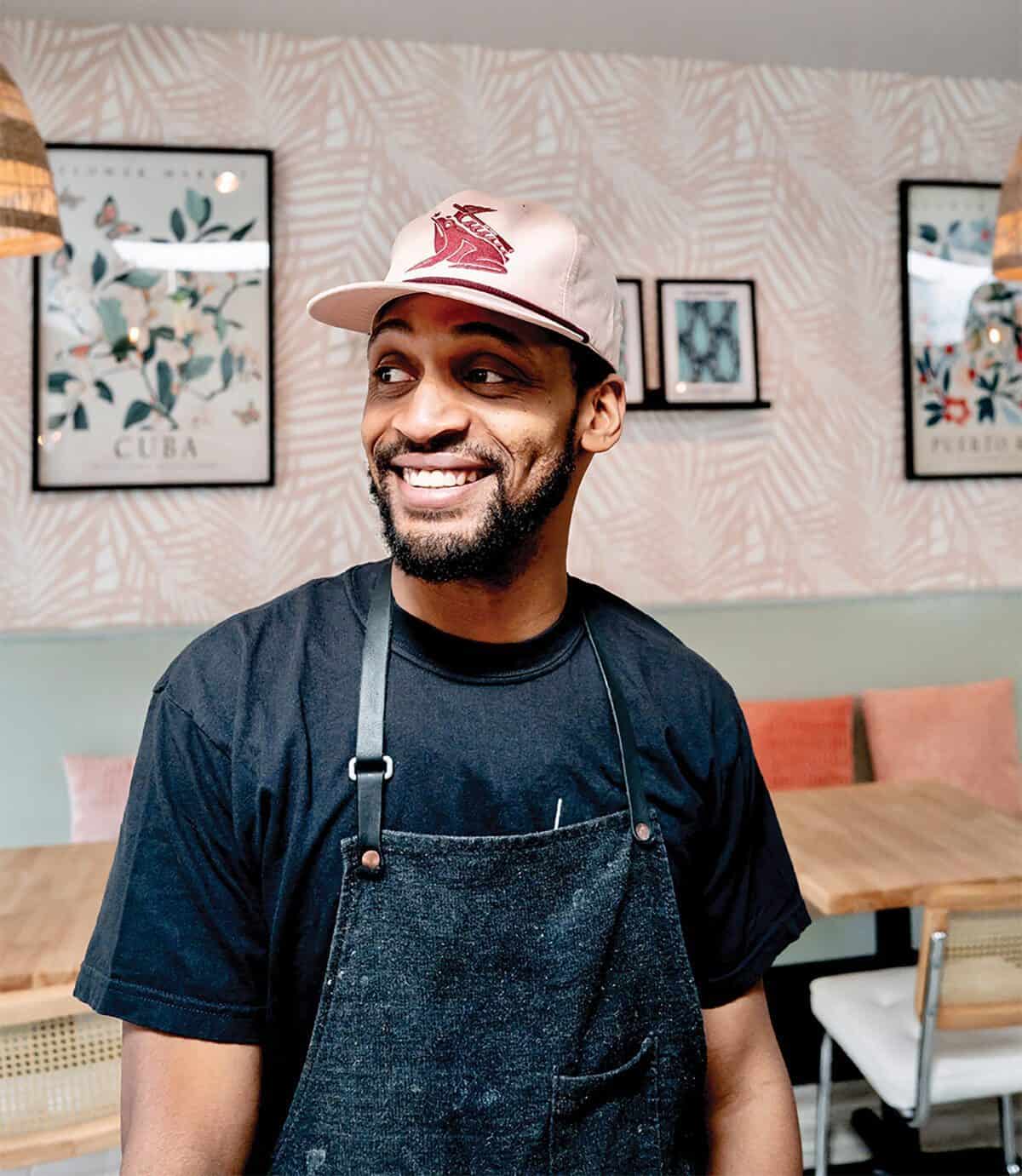 Johnny Reyes stands smiling in a modern Lenox cafe, wearing a black t-shirt, dark apron, and light pink cap, with framed artwork on the wall behind him.