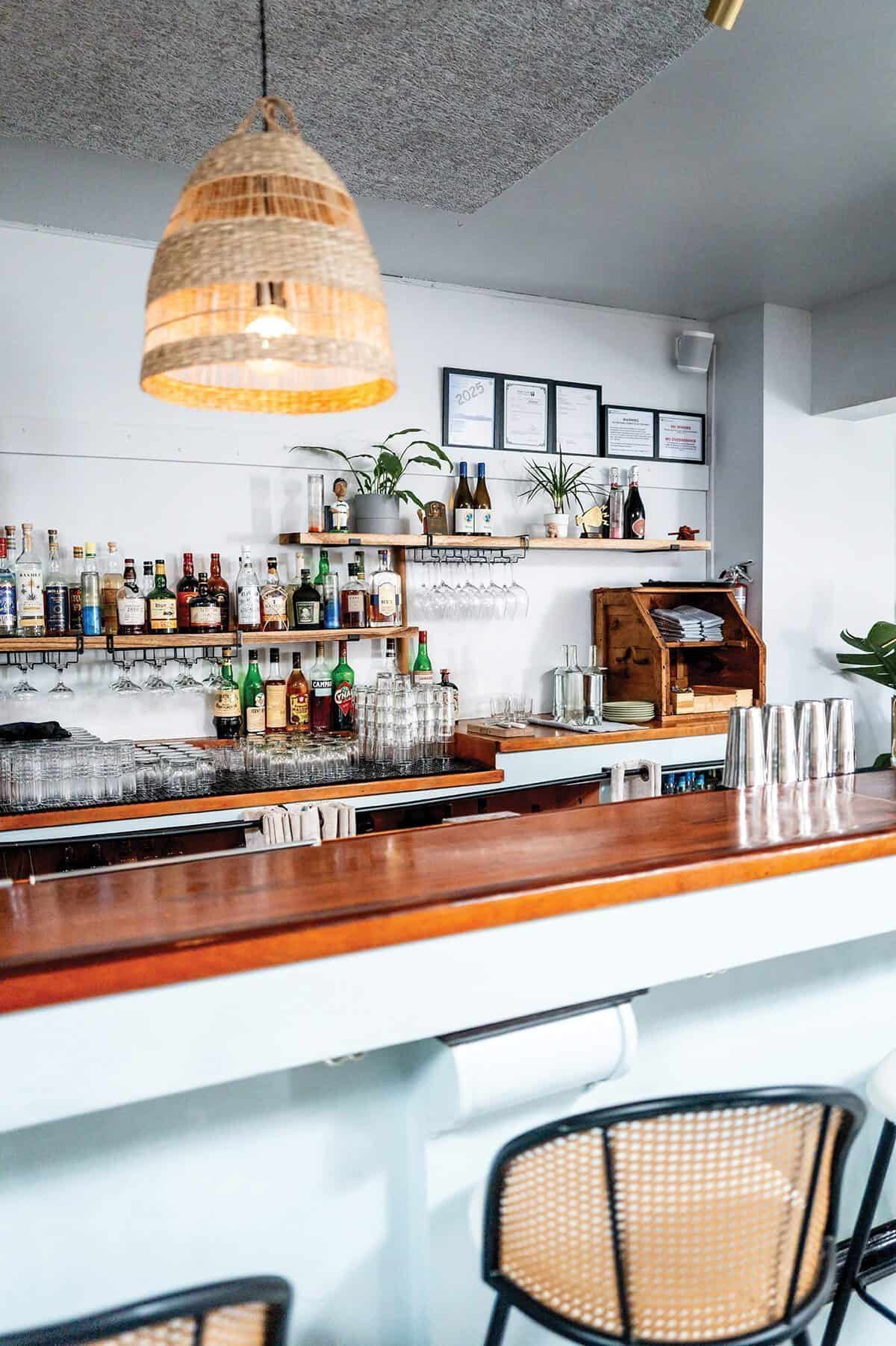 A modern Lenox bar with a wooden countertop, shelves of liquor bottles, glassware, a hanging wicker light fixture, and framed certificates on the wall curated by Johnny Reyes.