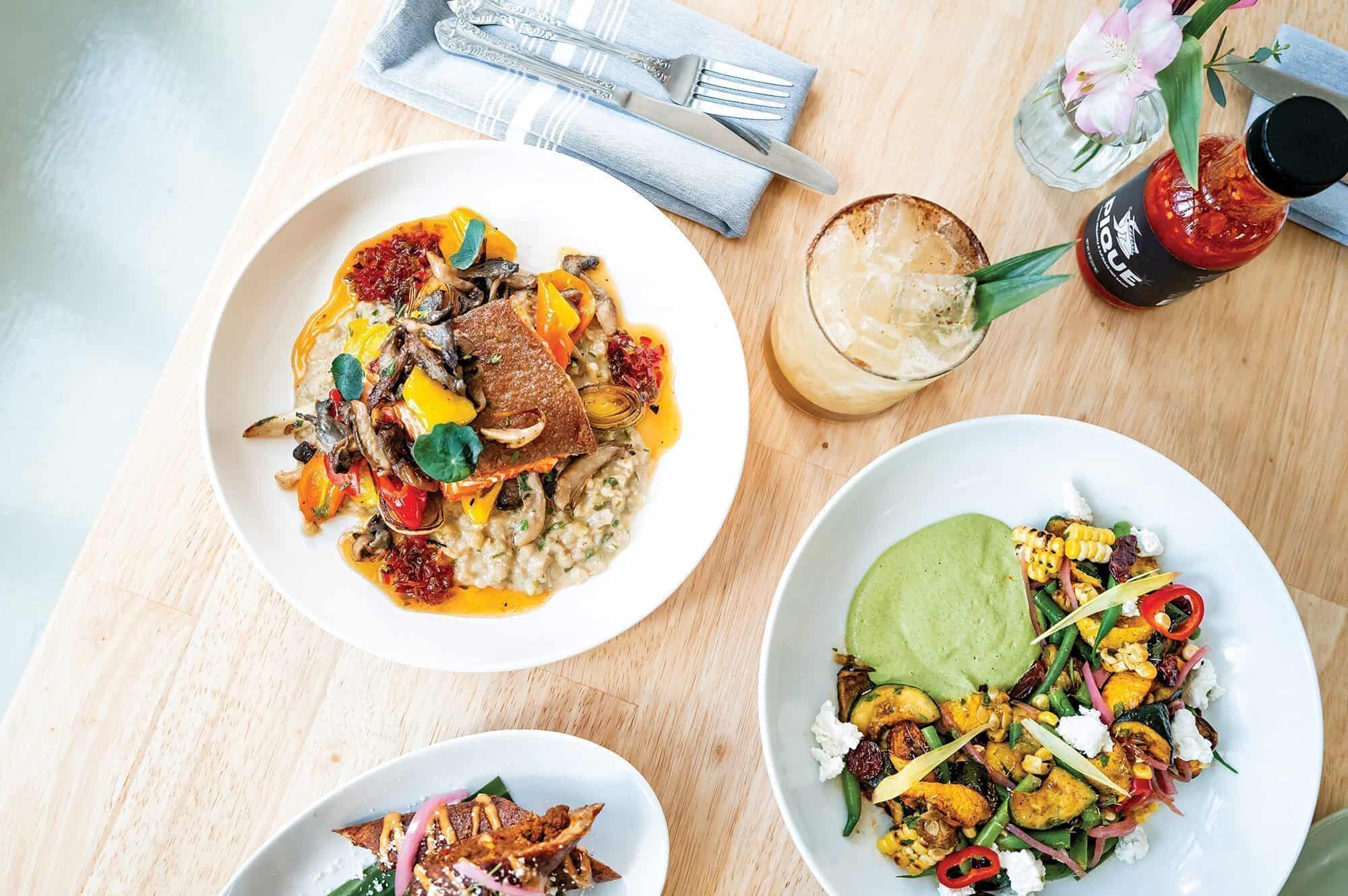 Three plated gourmet dishes by Johnny Reyes, a Lenox beverage with ice and garnish, cutlery, napkin, hot sauce, and a flower are artfully arranged on a light wooden table, viewed from above.