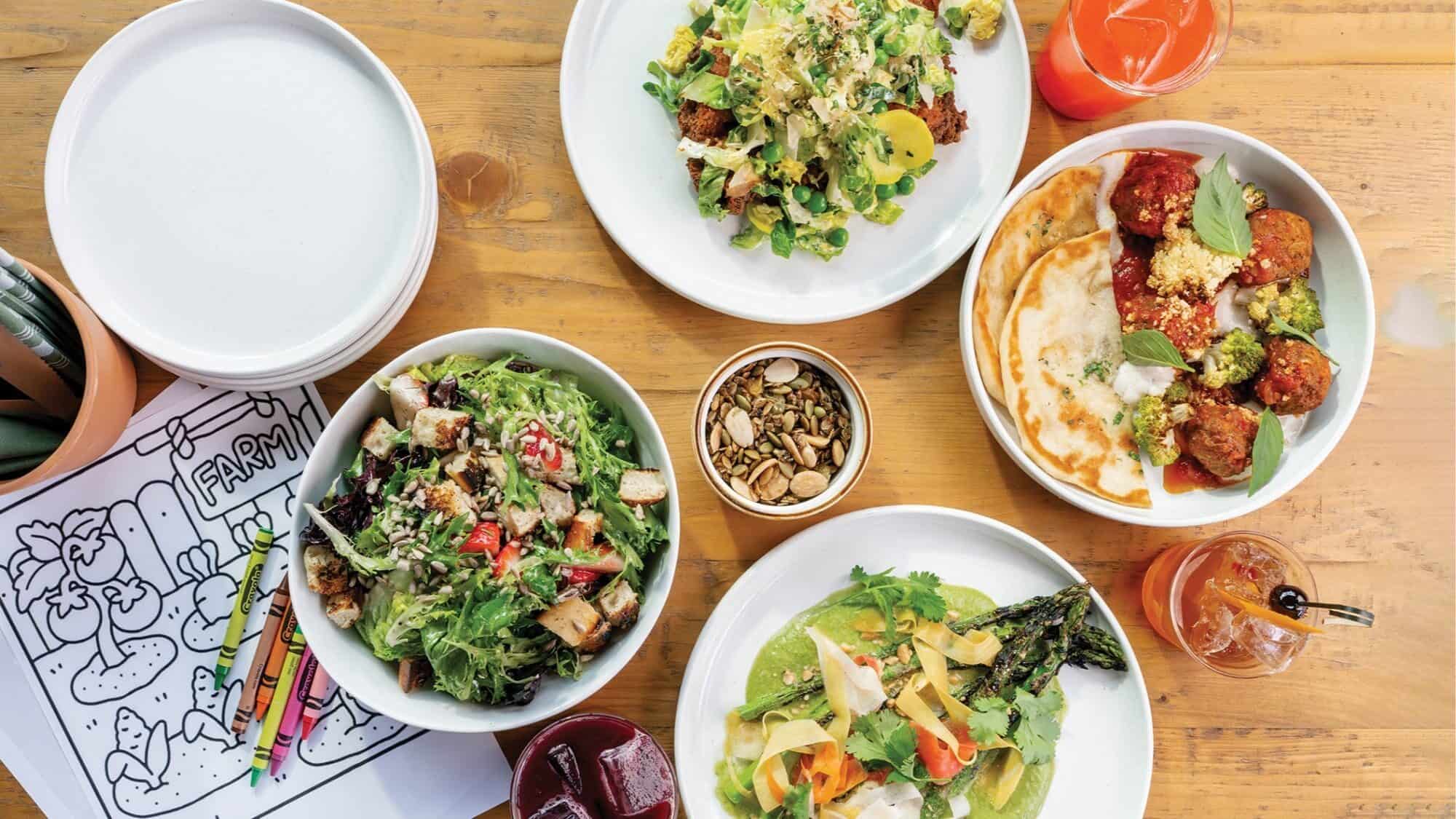 Overhead view of assorted salads made with locally sourced ingredients, a dish with meatballs and flatbread, two drinks, a cup of seeds, and a coloring sheet with crayons on a wooden table.