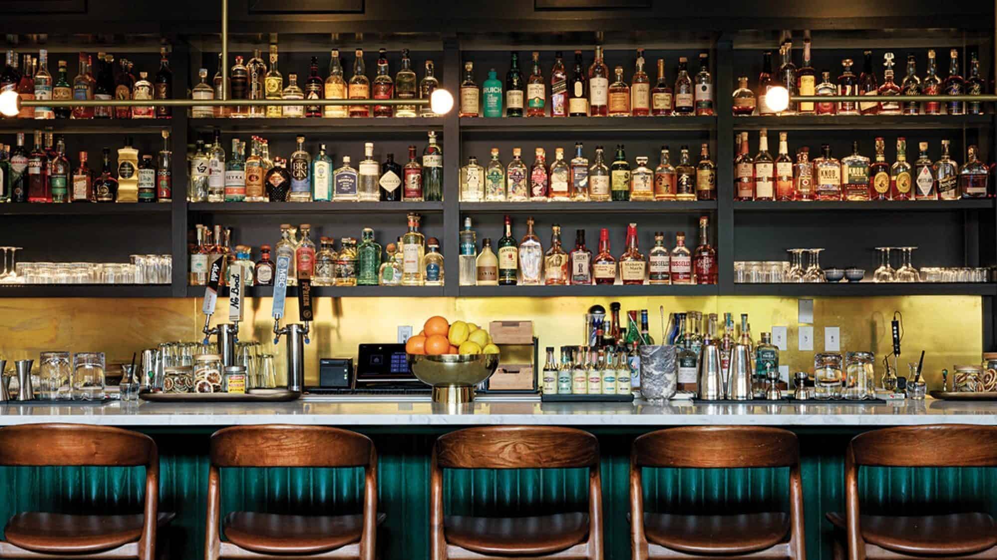 A bar interior at Ramie, one of the standout Capitol Hill restaurants, features liquor bottles on shelves, a countertop with bar tools and fruit, and four wooden bar stools inviting guests to enjoy Trinh Nguyen’s signature creations.