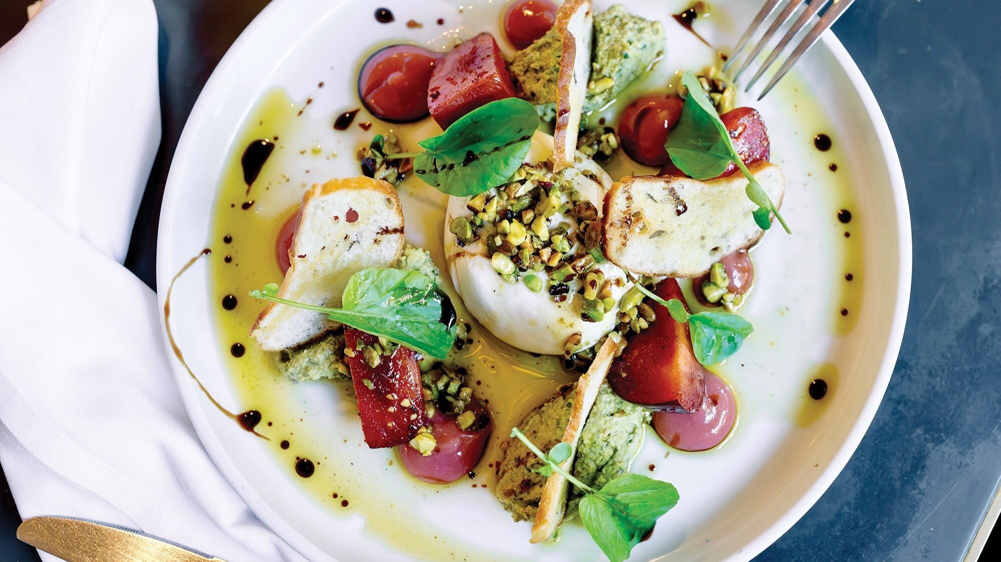 A plated dish with burrata cheese, roasted beets, pesto, toasted bread slices, microgreens, chopped nuts, and balsamic glaze masterfully arranged on a white plate—a true masterclass in flavor and presentation.