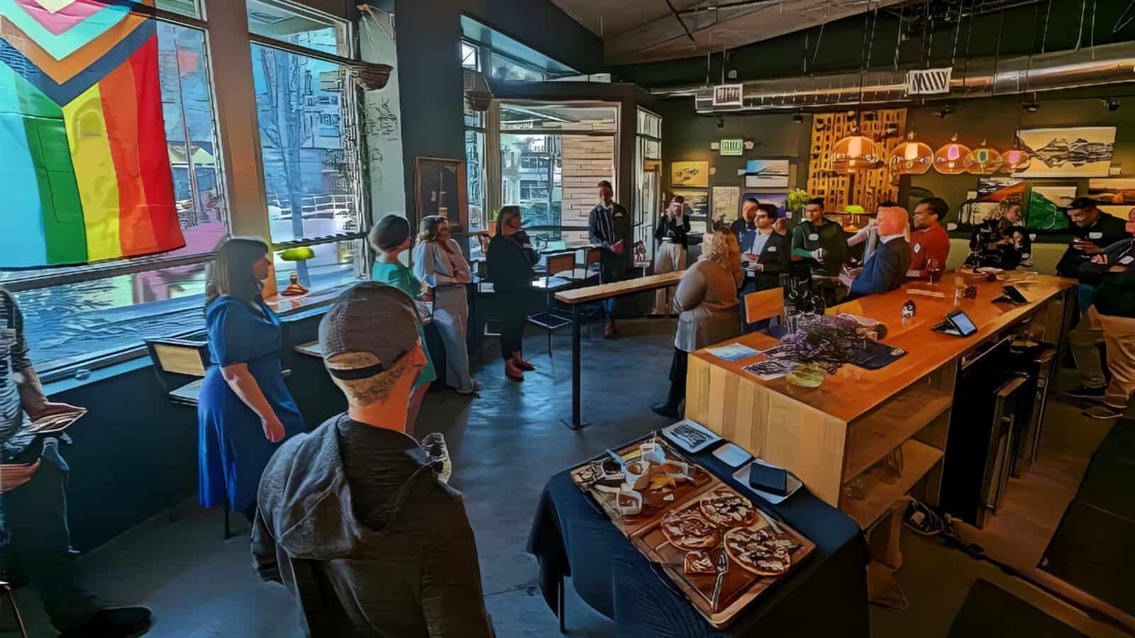 A group of people gather in a modern cafe space with a large pride flag, snacks on the table, and art on the walls as vibrant celebrations honor Seattle’s rich LGBTQIA+ history.