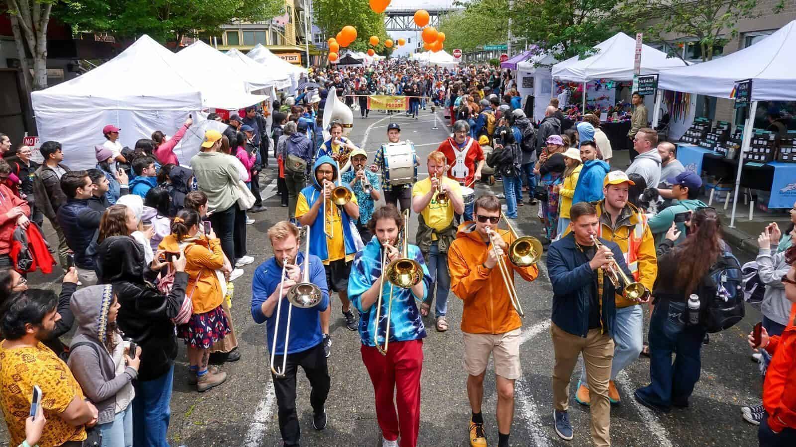 A brass band marches through a crowded outdoor street festival lined with vendor tents, as people watch and take photos—one of the many fun things to do in Seattle near MOPOP and SeattleRep.