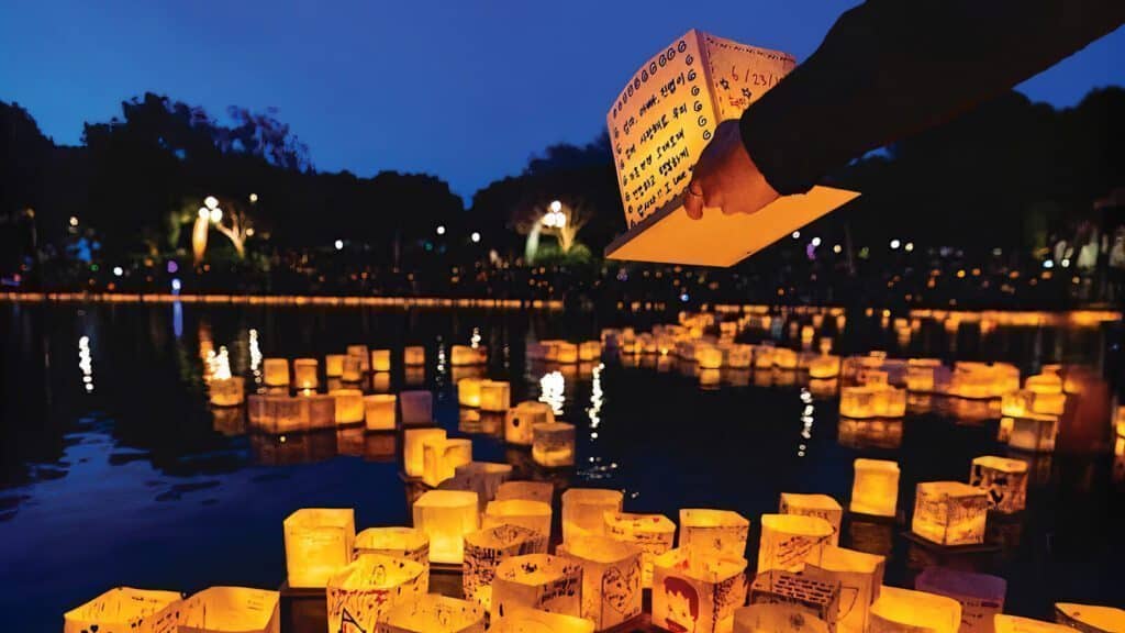 A hand releases a lantern with handwritten messages onto water, joining many illuminated lanterns floating on a lake during dusk—a serene scene reminiscent of art and food during the Impressionism era.