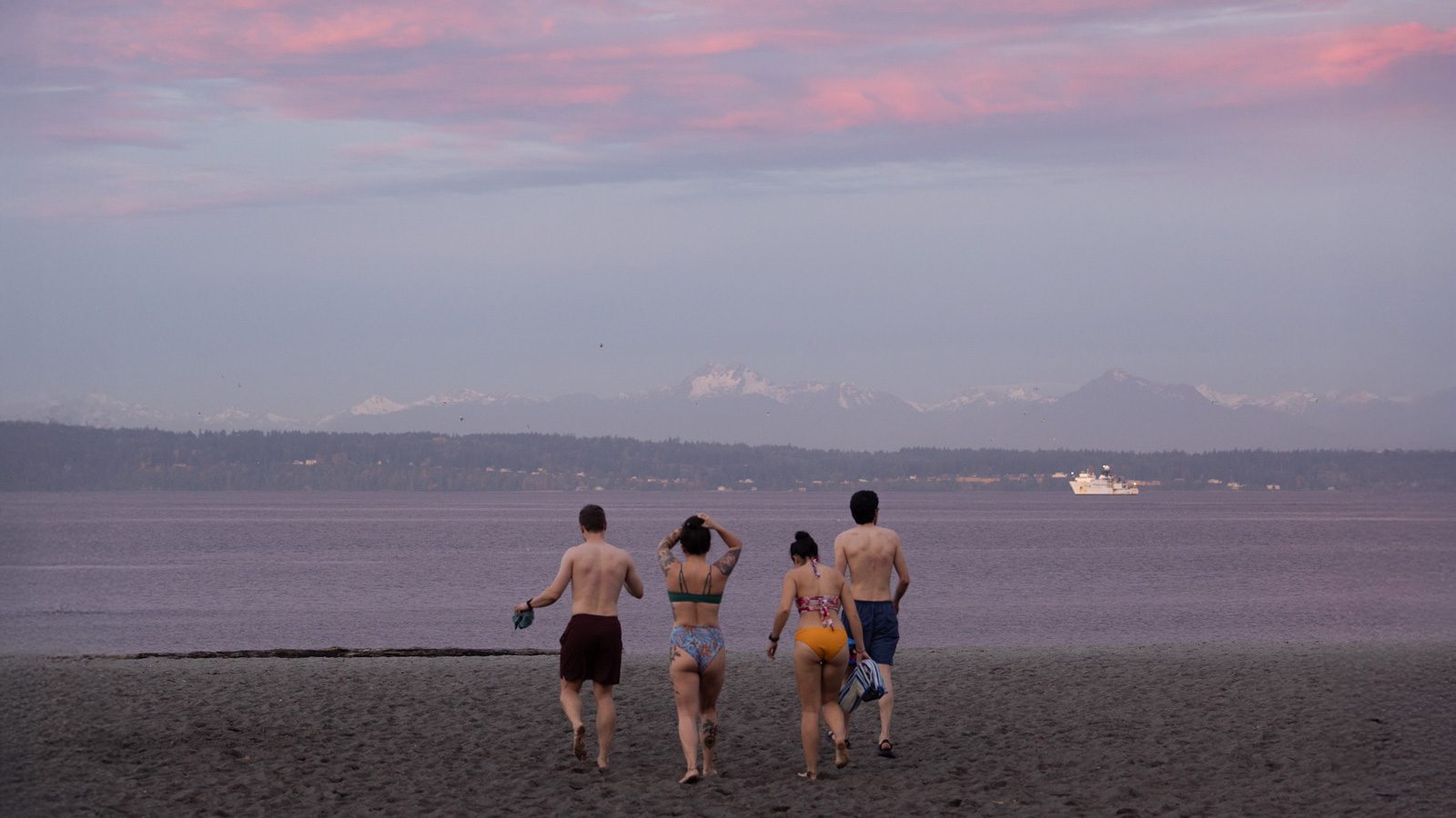 Four people in swimwear walk toward the water on a Seattle beach at sunset, with mountains and a ferry in the background. It's a New Year for all types, embracing the chill and beauty of the moment.