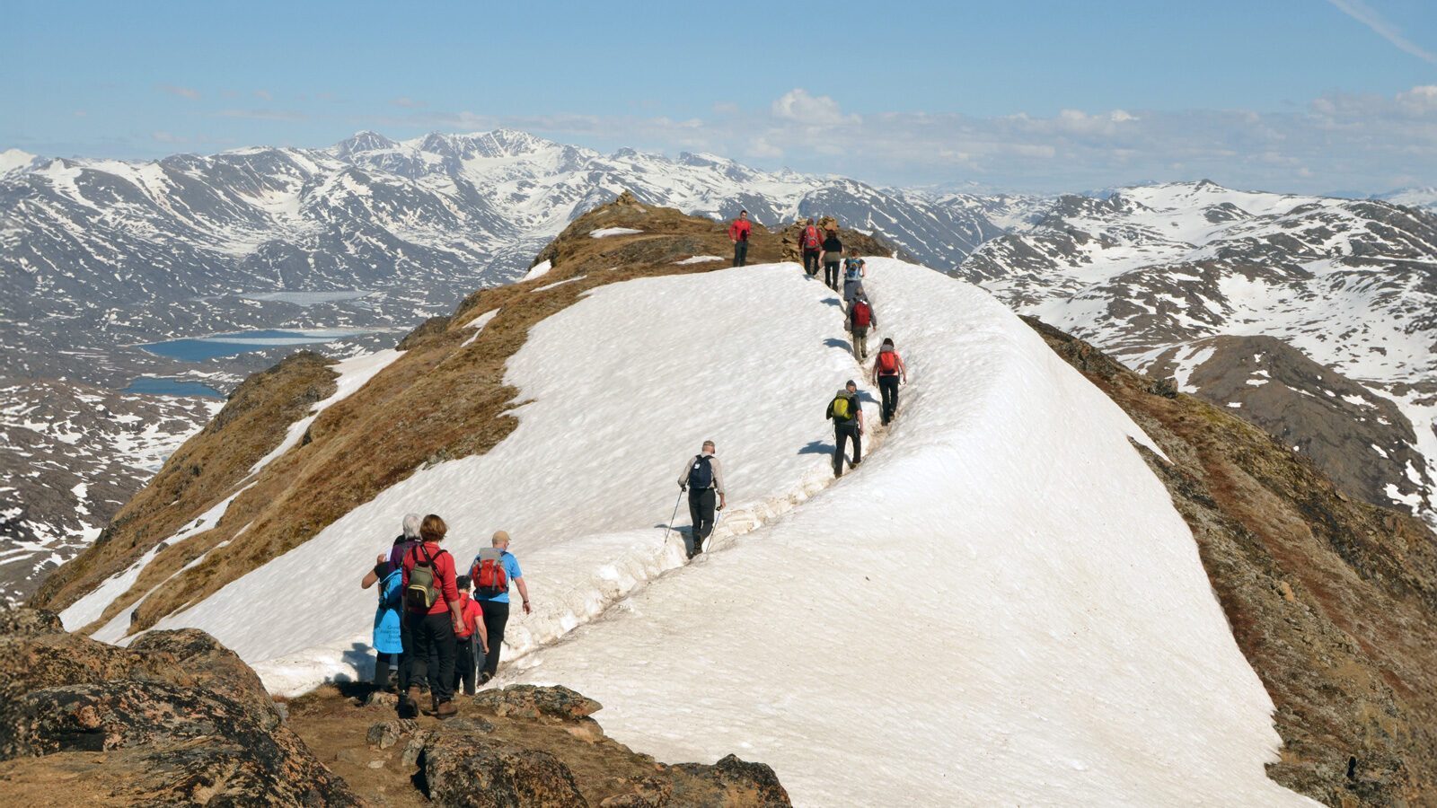 A group of hikers walk along a snowy mountain ridge in Greenland, surrounded by rugged, snow-capped peaks and distant icebergs under a partly cloudy sky.