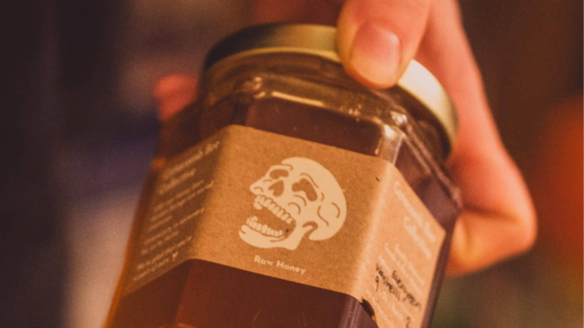 A hand holds a glass jar of raw honey with a brown label featuring an illustration of a skull.