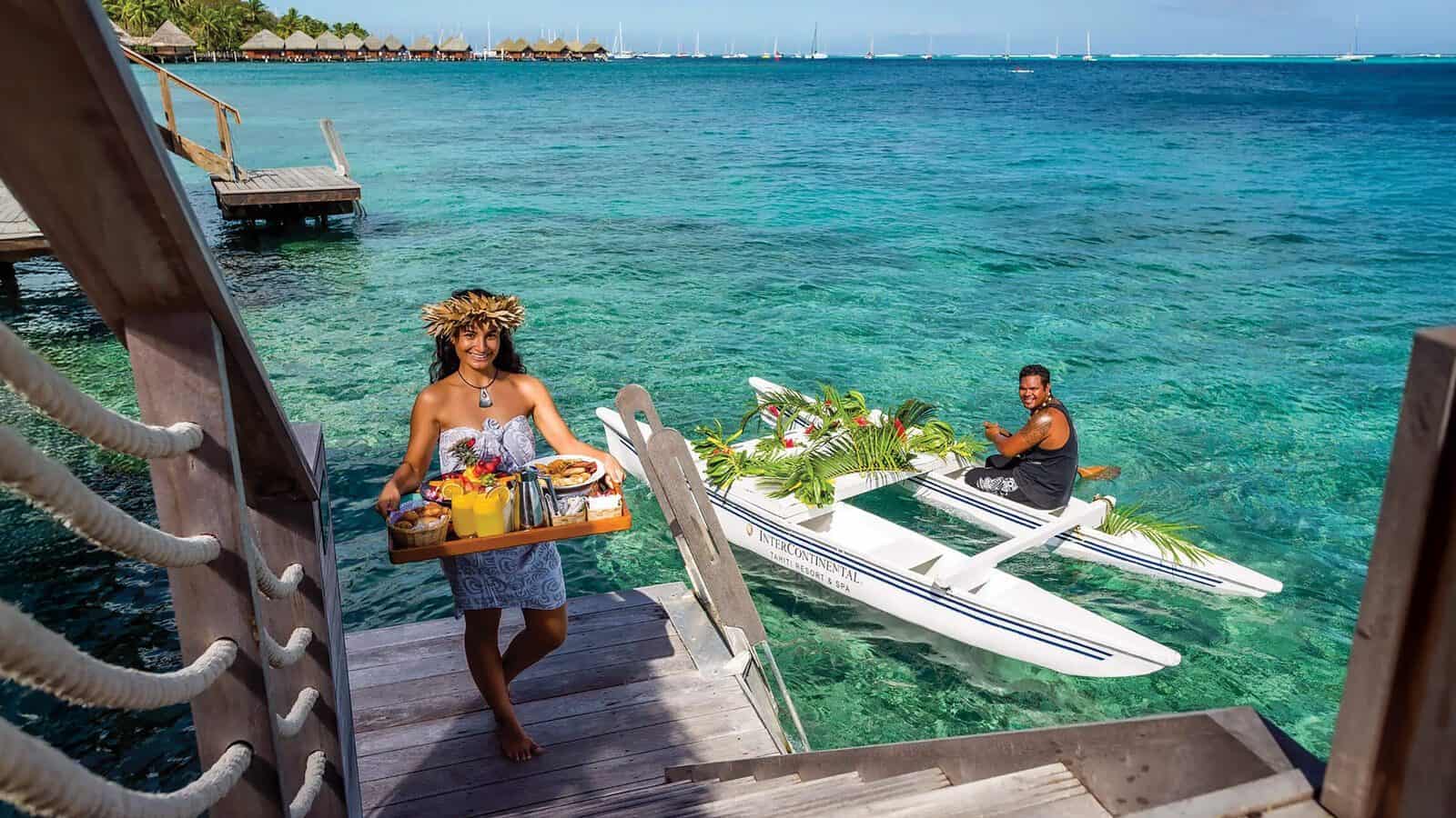 A woman in traditional attire carries a breakfast tray up wooden steps from the water in Tahiti, while a man sits in an outrigger canoe on the clear blue lagoon.