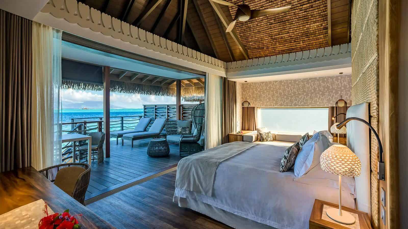 A modern Tahiti overwater bungalow bedroom with a large bed, wooden ceiling, and an open view to a deck with lounge chairs overlooking the ocean.