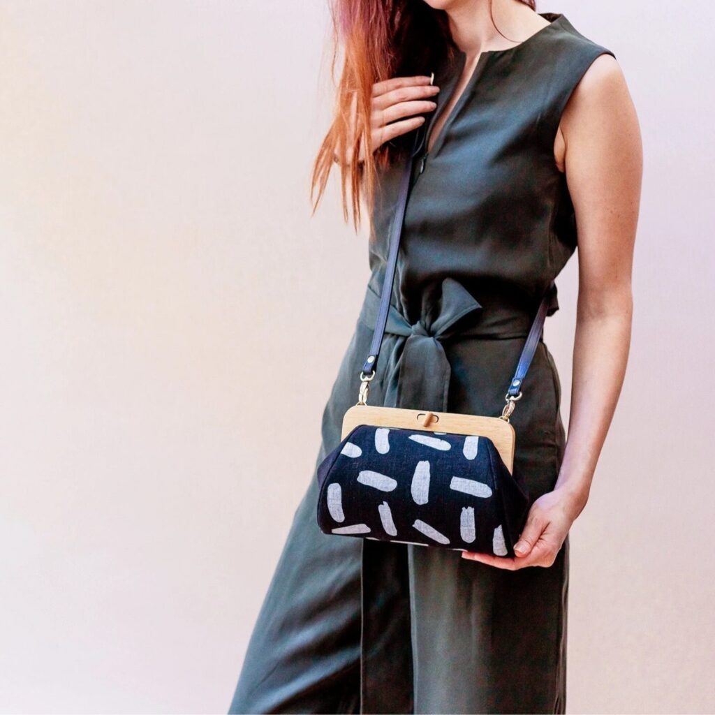 A woman in a sleeveless dark green jumpsuit holds a black purse with a tan frame and abstract light gray patterns.