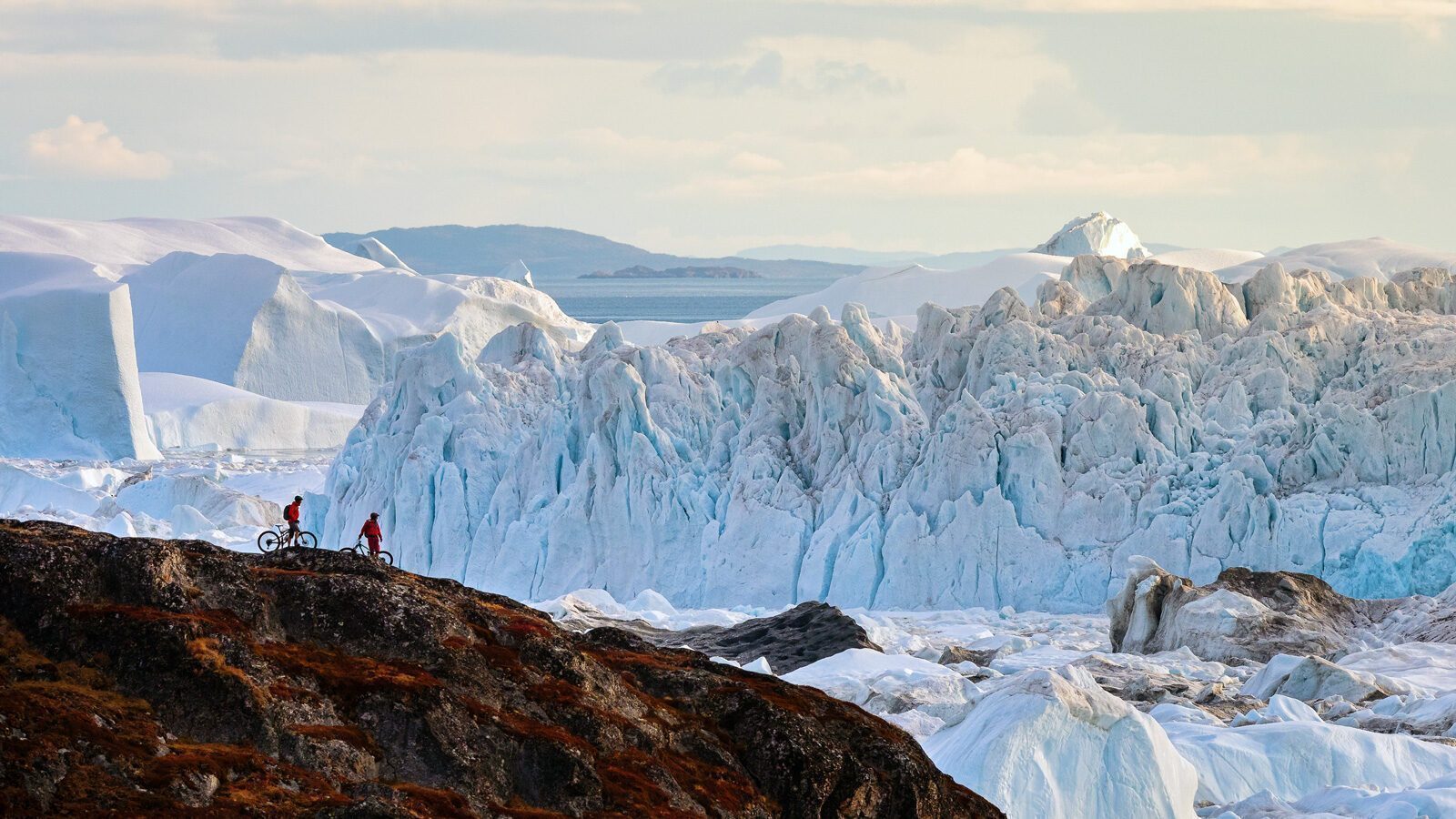 Two cyclists ride along a rocky ridge in Greenland, with large icebergs and glaciers in the background under a partly cloudy sky—an awe-inspiring scene perfect for adventurous expeditions.